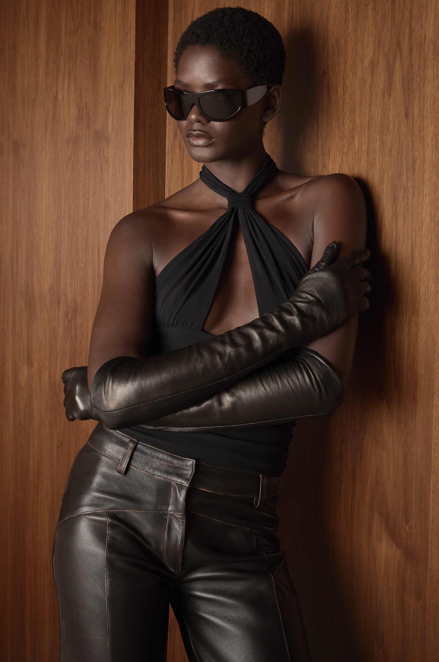Person wearing sunglasses, a black halter top, leather gloves, and leather pants against a wooden wall.