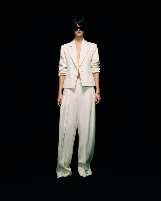 Model wearing a cream blazer with ruched sleeves worn open over wide-leg cream trousers, with dark sunglasses, full-length front view against a black background