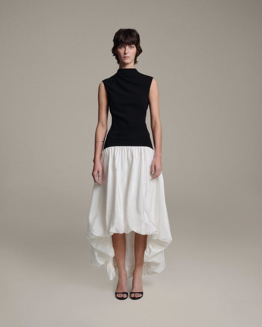 Model wearing a black sleeveless draped mock-neck top tucked into a white bubble-hem midi skirt with a high-low hem, full-length front view against a taupe background