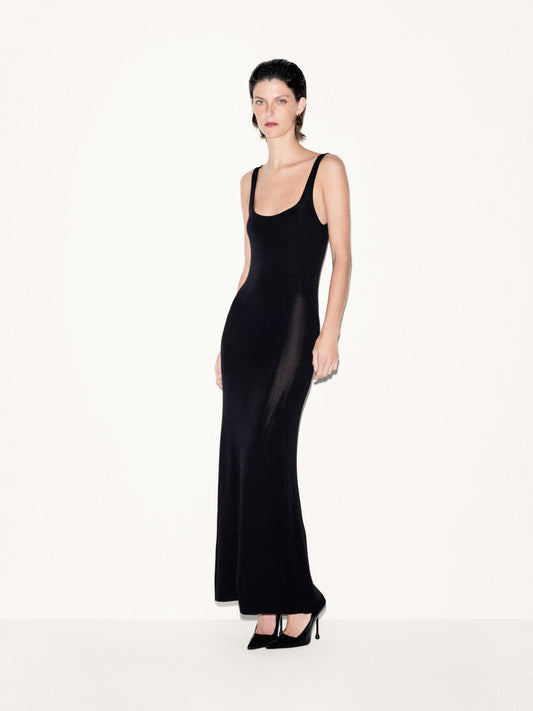 A model wearing a black sleeveless maxi dress with sheer mesh side panels and black pointed-toe heels, standing against a plain off-white studio background.