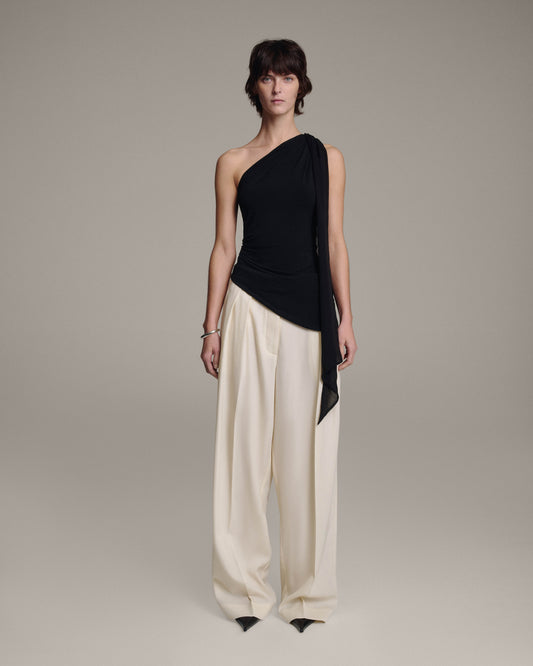Model wearing a black one-shoulder top with a sheer chiffon shoulder tie draping down one side, paired with wide-leg cream trousers, full-length front view against a taupe background