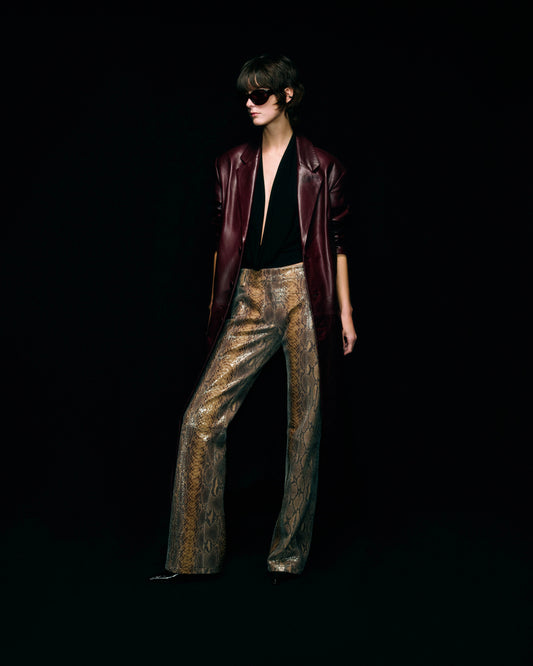 A model stands against a solid black background wearing a dark burgundy leather jacket, a black halter top, and snakeskin patterned leather trousers.