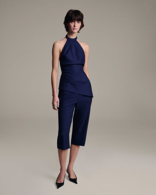 A model wearing a navy blue sleeveless halter top and matching cropped capri pants, paired with black pointed-toe heels, against a solid warm gray studio background.
