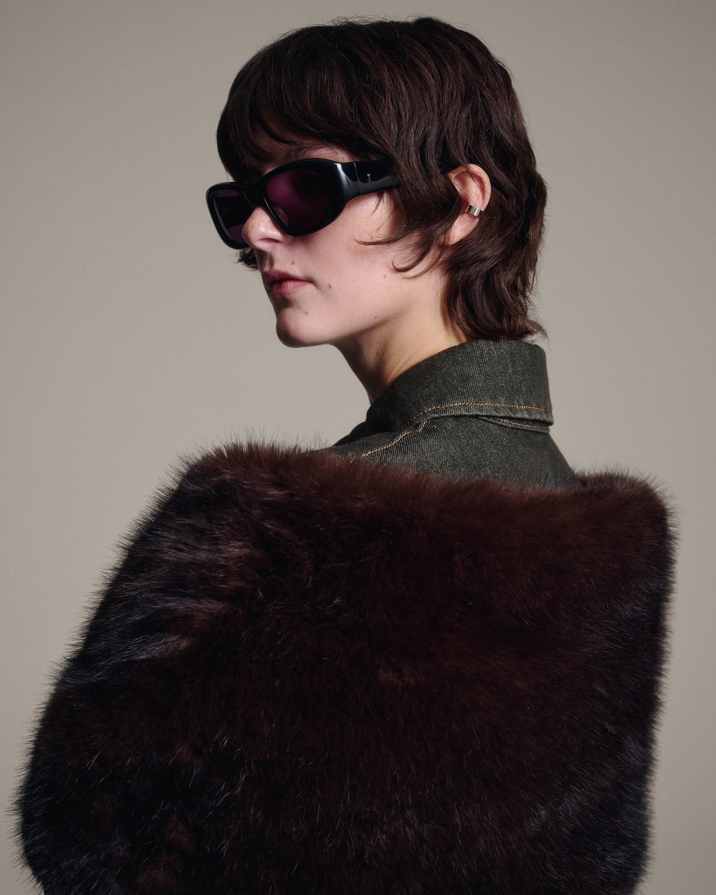 A model with short dark hair wearing black sunglasses and a dark brown fur stole over a dark denim jacket, photographed against a solid beige background.
