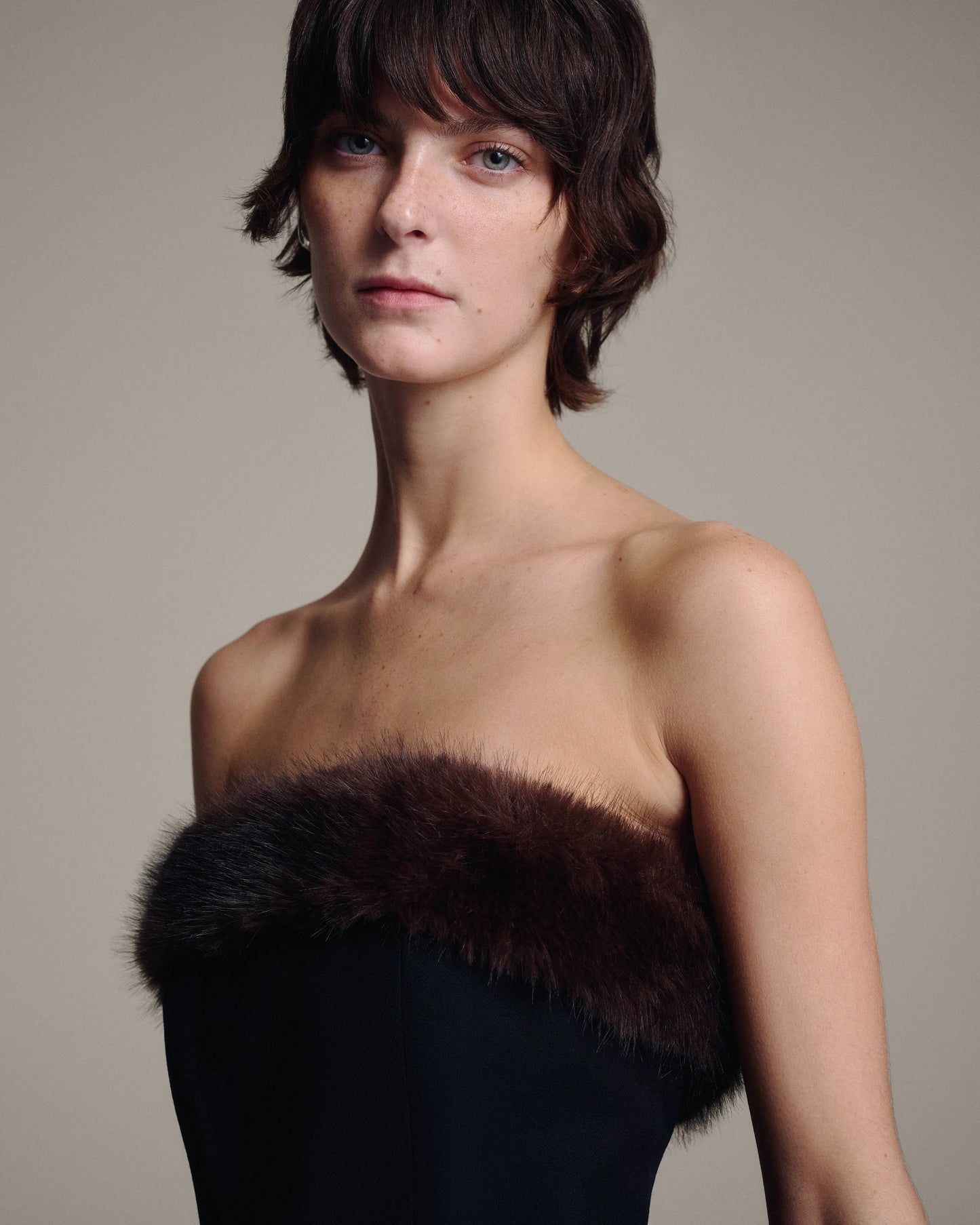 A model with short dark hair wearing a strapless black dress with a dark brown fur trim, photographed against a plain beige studio background.