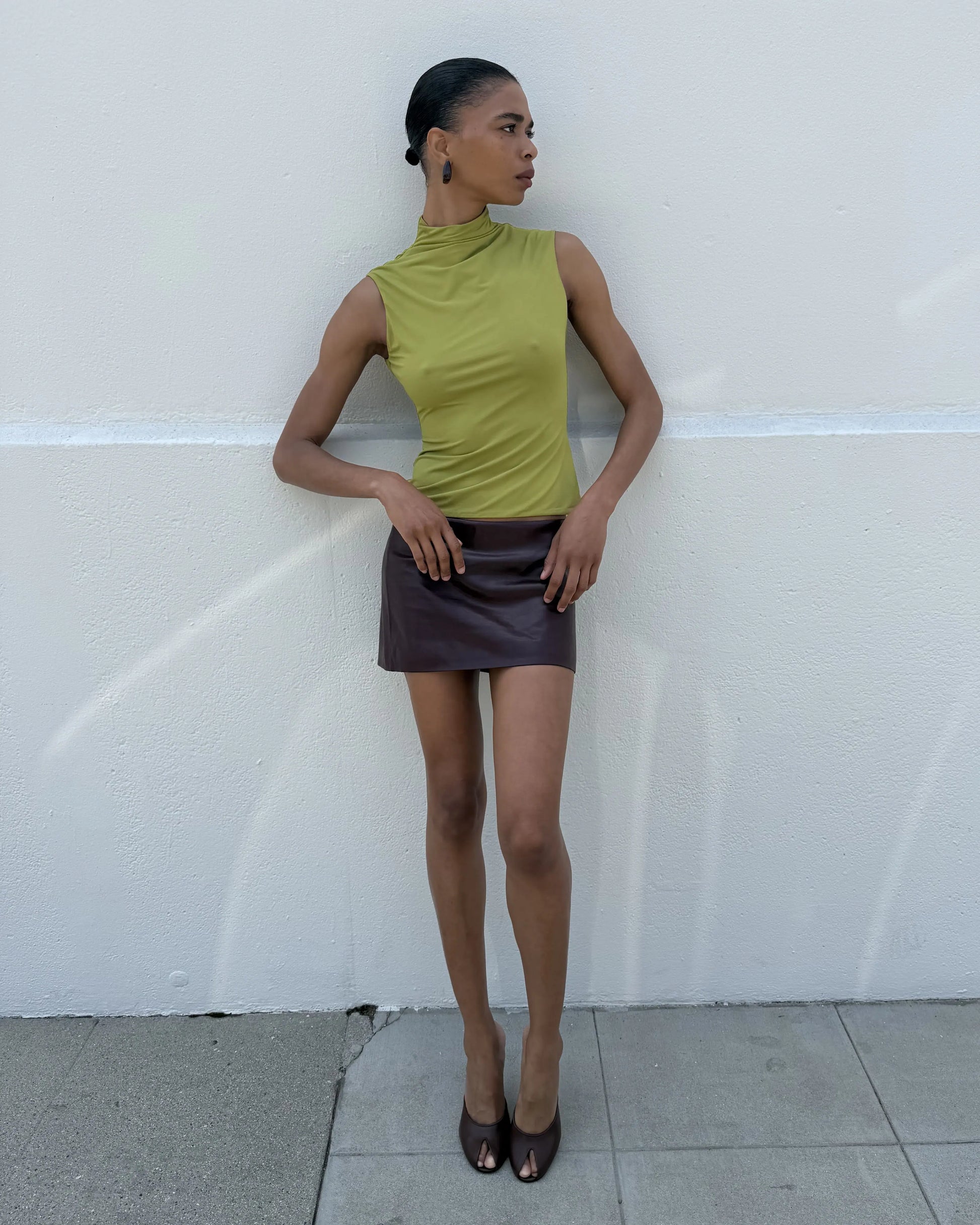 "Model wearing an olive green sleeveless mock-neck top with a draped high neck and a deep brown leather mini skirt with deep brown peep-toe heels, full-length front view against a white wall