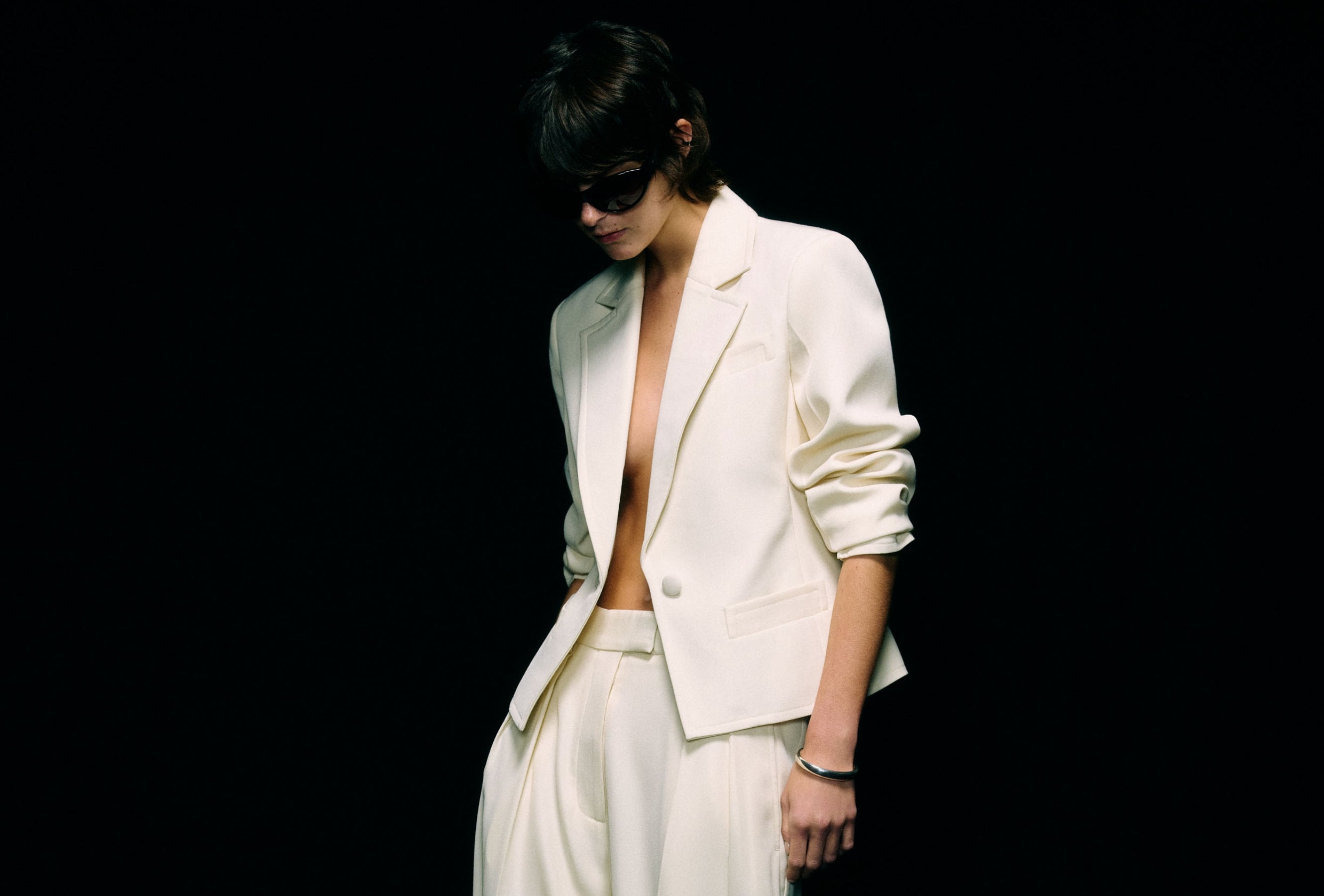 Model wearing a cream blazer worn open over wide-leg cream trousers, full-length front view against a black background
