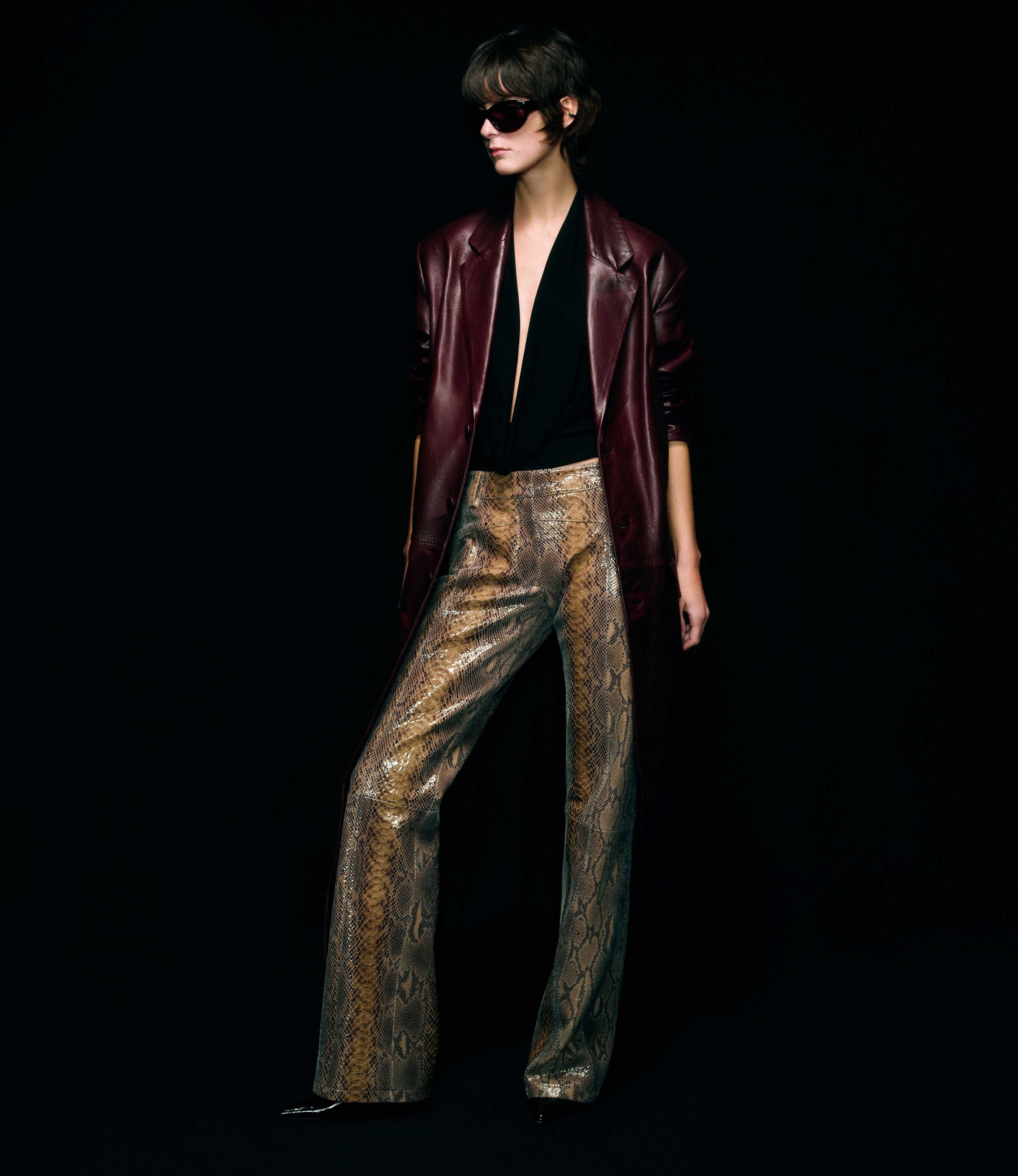 A fashion model wearing a dark burgundy leather blazer, a black top, and snakeskin-patterned leather trousers, set against a solid black background.