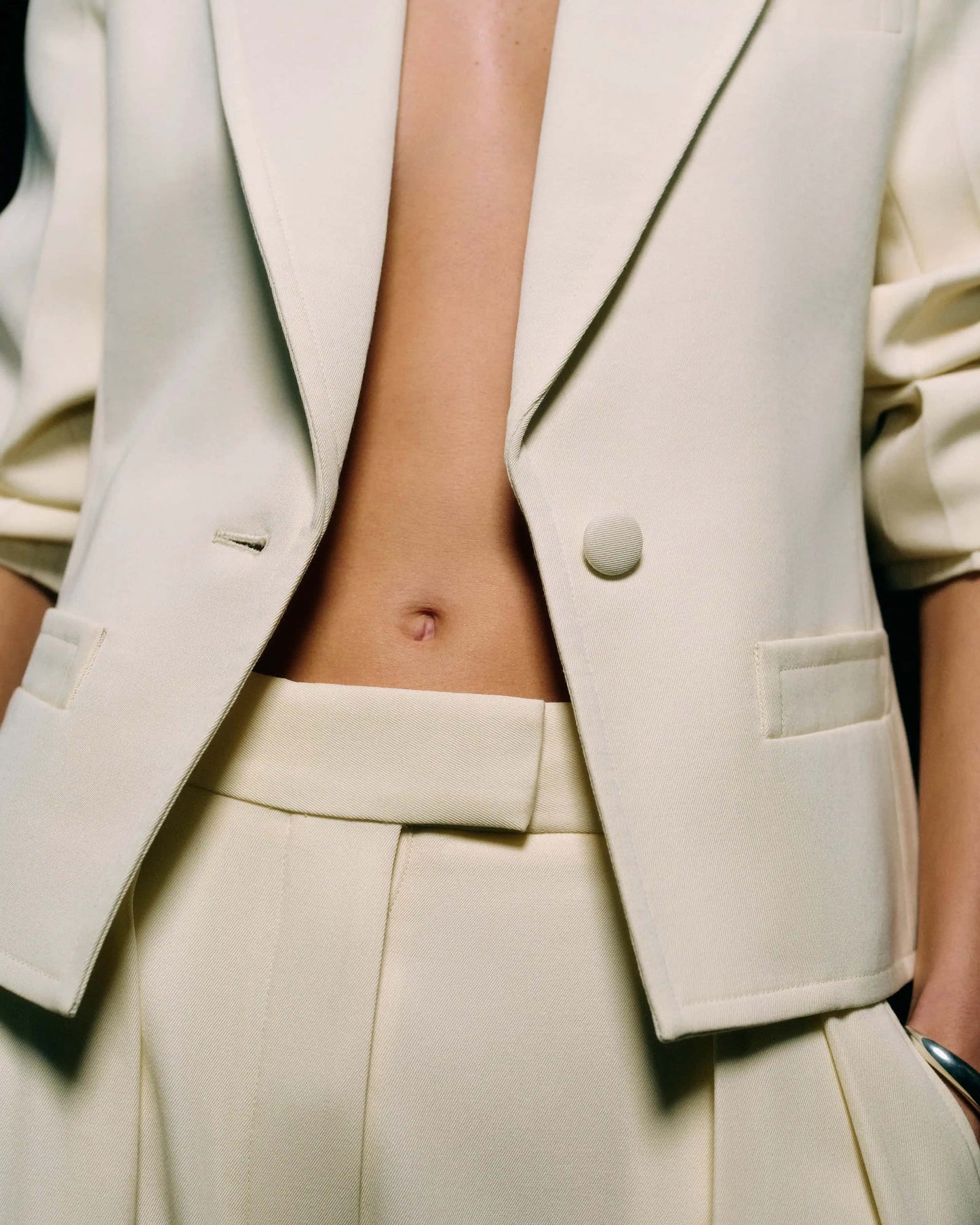 Close-up of the cropped front hem and single-button closure of a cream tailored blazer over the waistband of cream trousers against a dark background