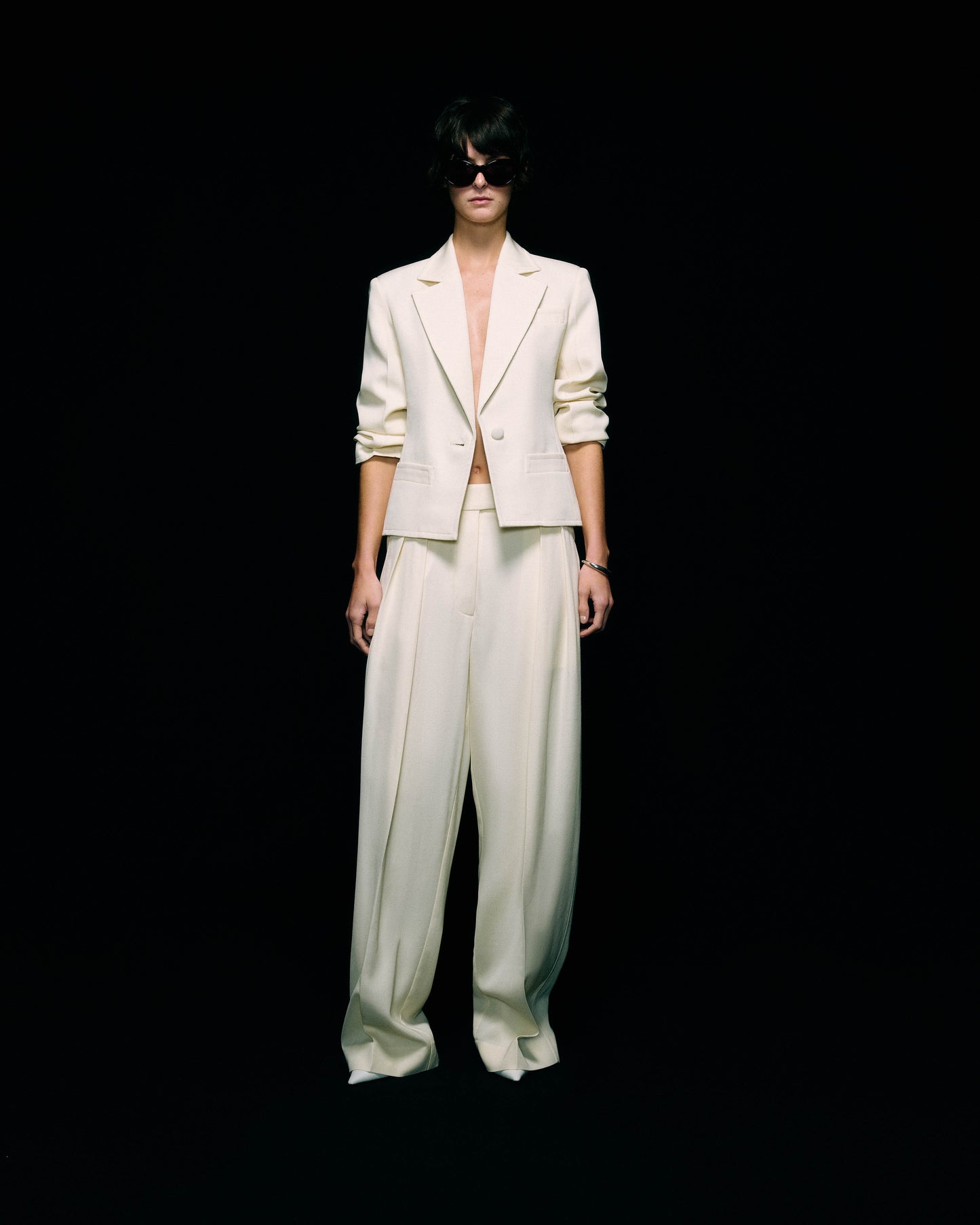 Model wearing a cream blazer with ruched sleeves worn open over wide-leg cream trousers, with dark sunglasses, full-length front view against a black background