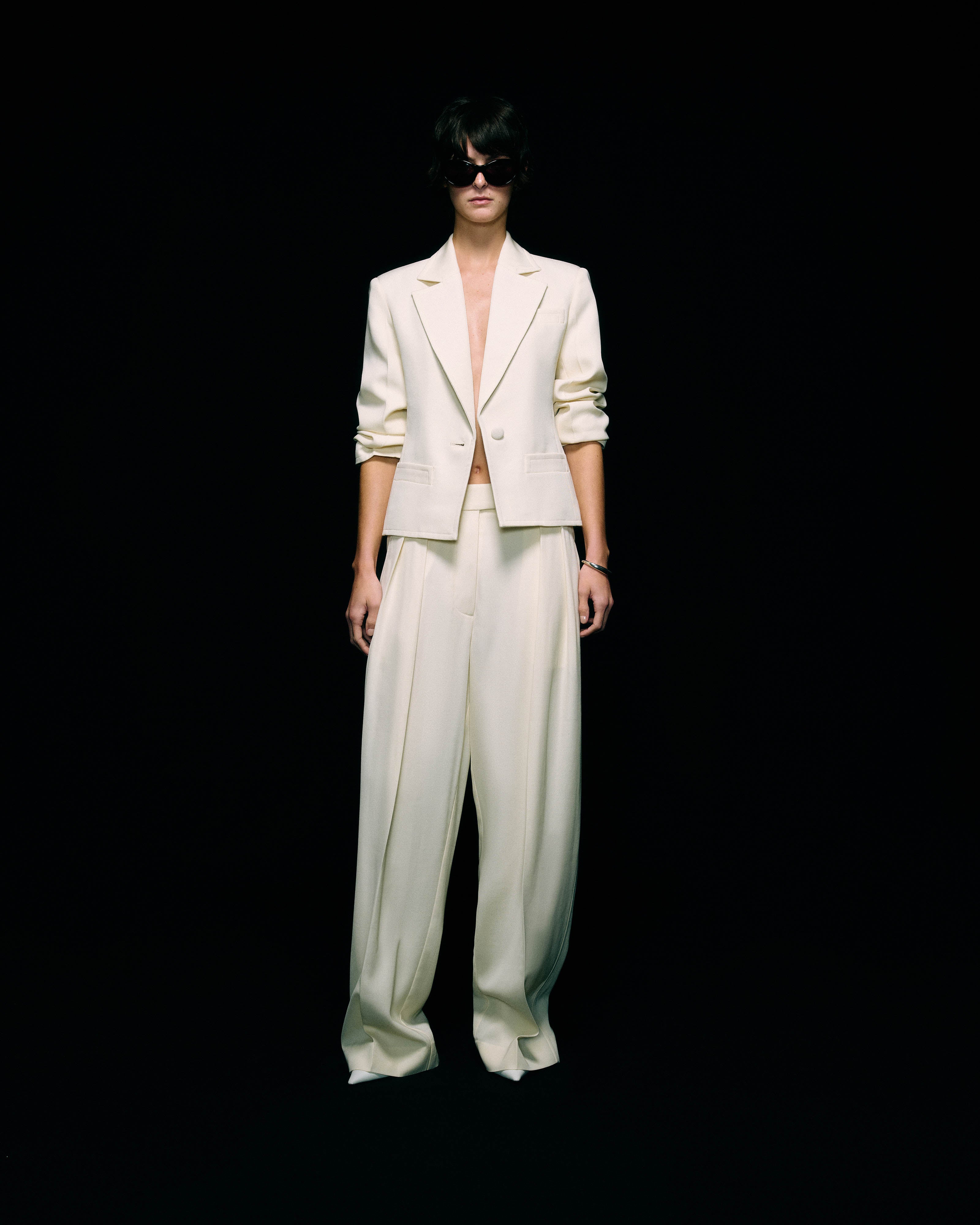 Model wearing a cream blazer with ruched sleeves worn open over wide-leg cream trousers, with dark sunglasses, full-length front view against a black background