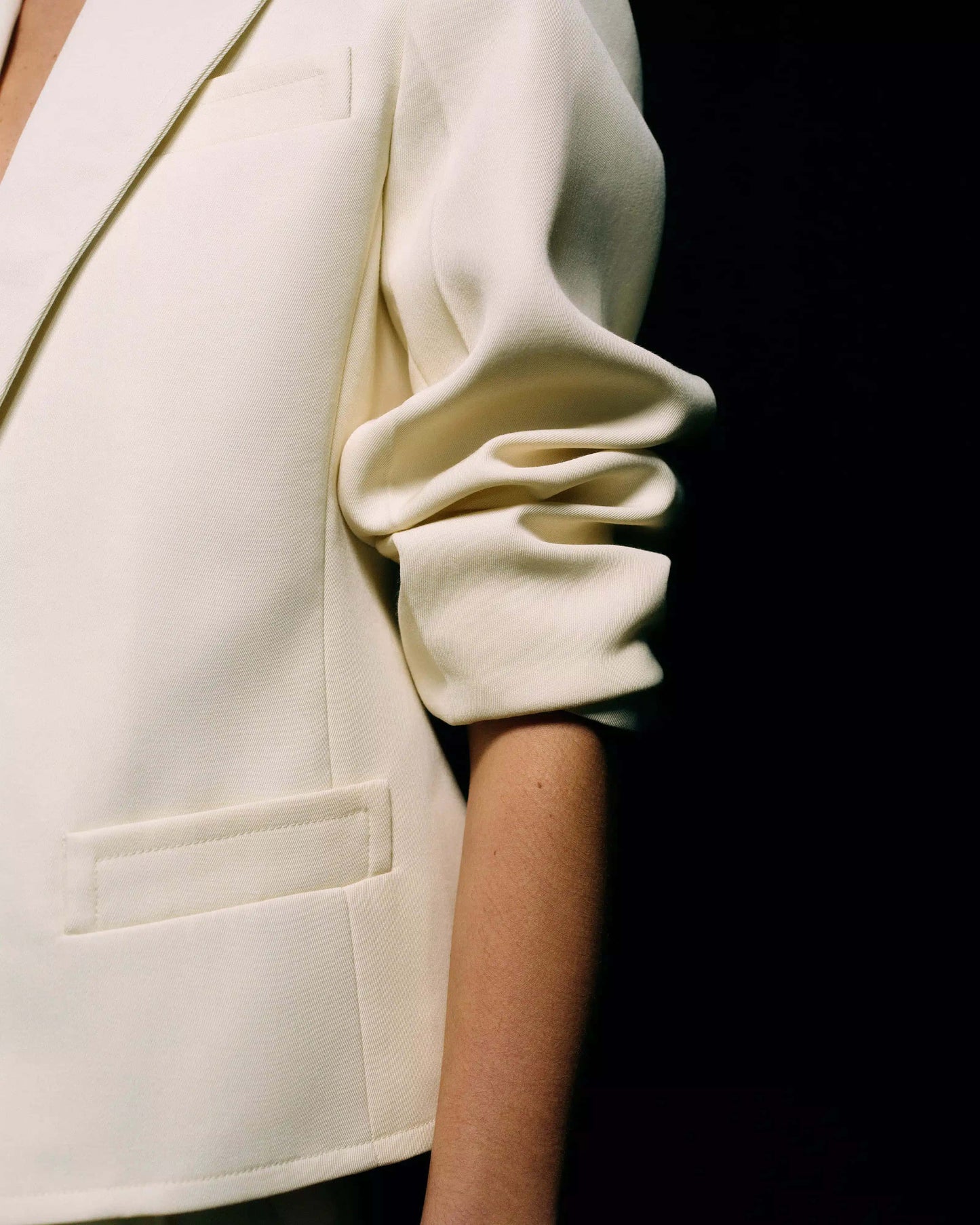 Close-up of the ruched gathered three-quarter sleeve and welt pocket on a cream tailored blazer against a black background