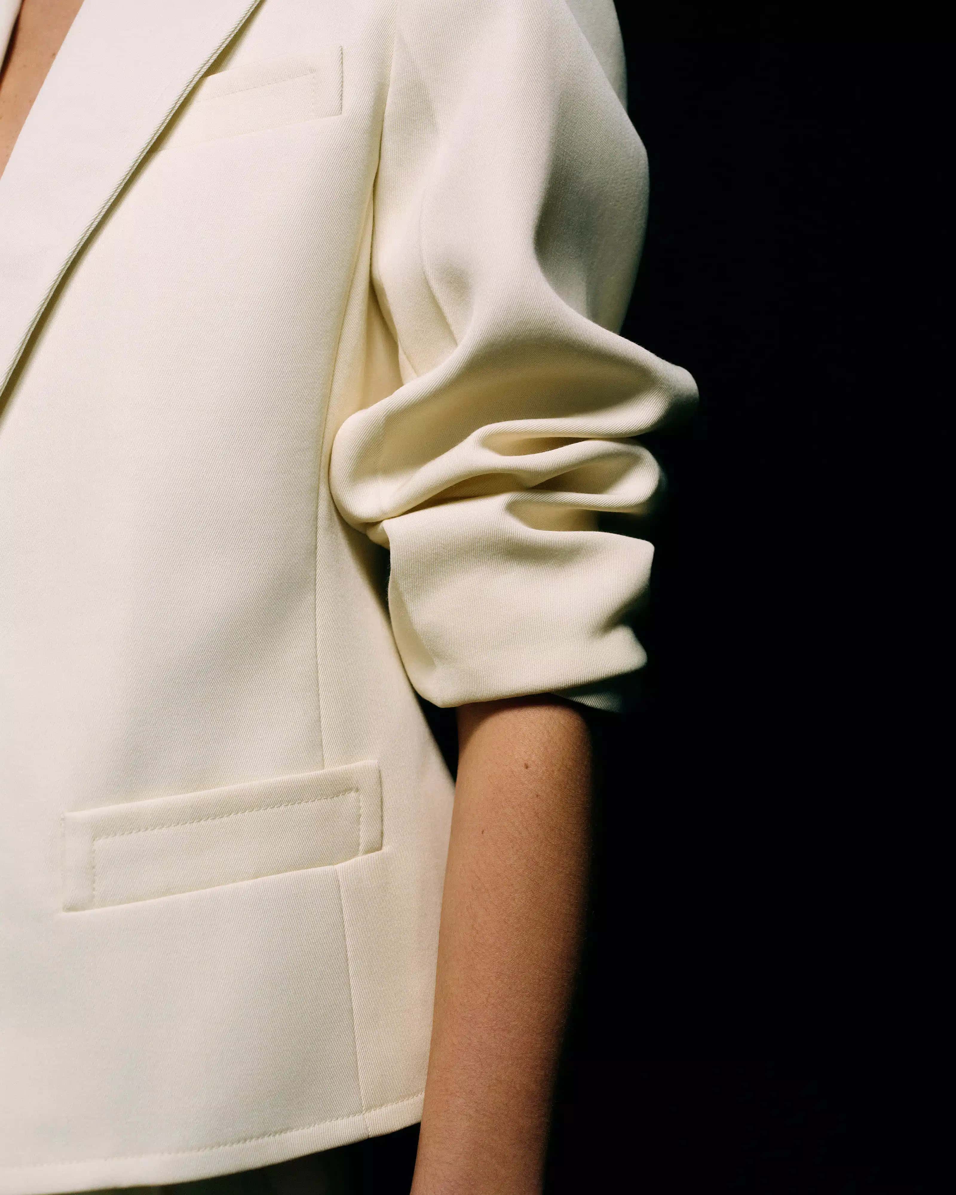 Close-up of the ruched gathered three-quarter sleeve and welt pocket on a cream tailored blazer against a black background