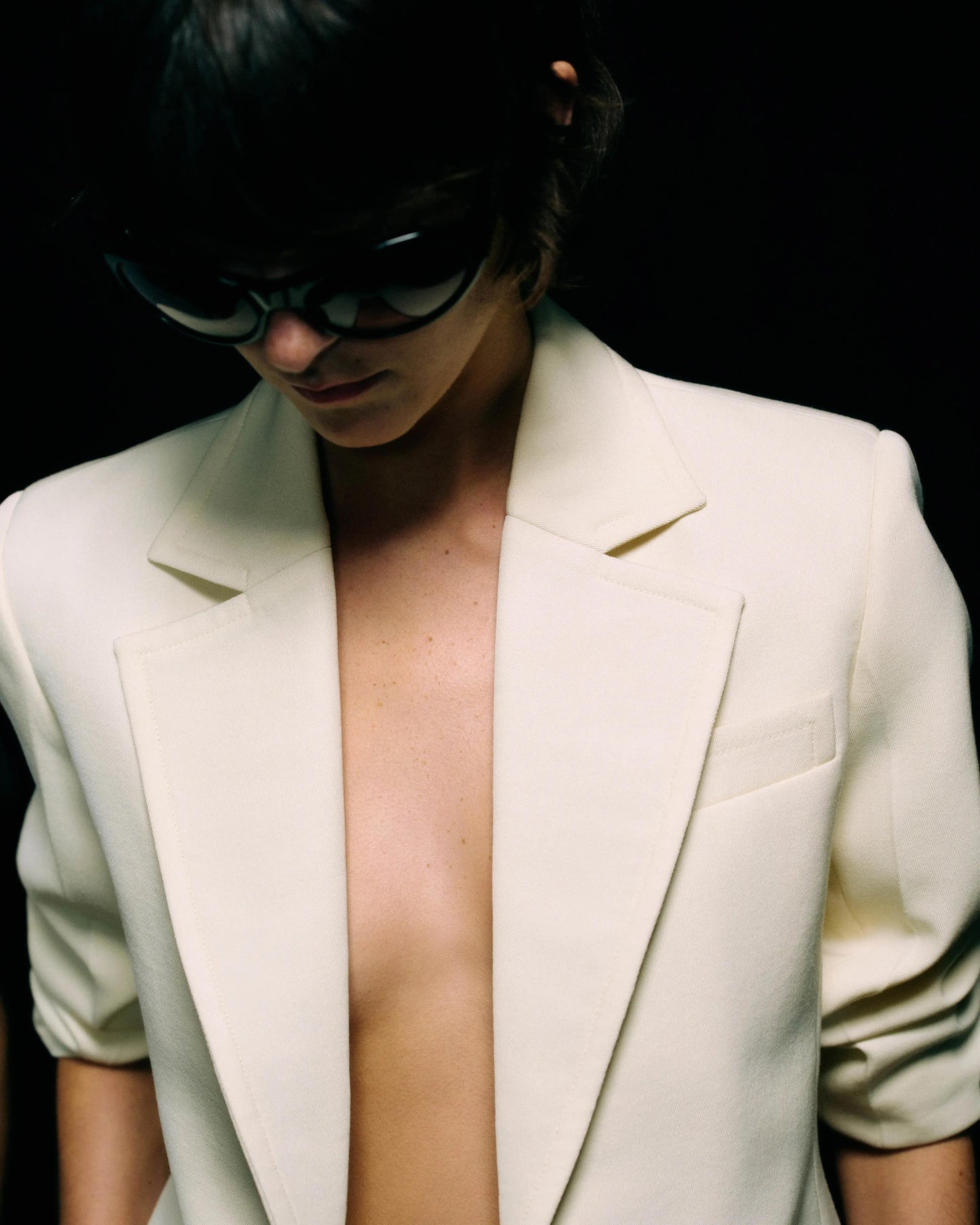 Model in a cream tailored blazer worn open over a bare chest with wide notch lapels and ruched three-quarter sleeves, dark sunglasses, upper-body crop against a black background