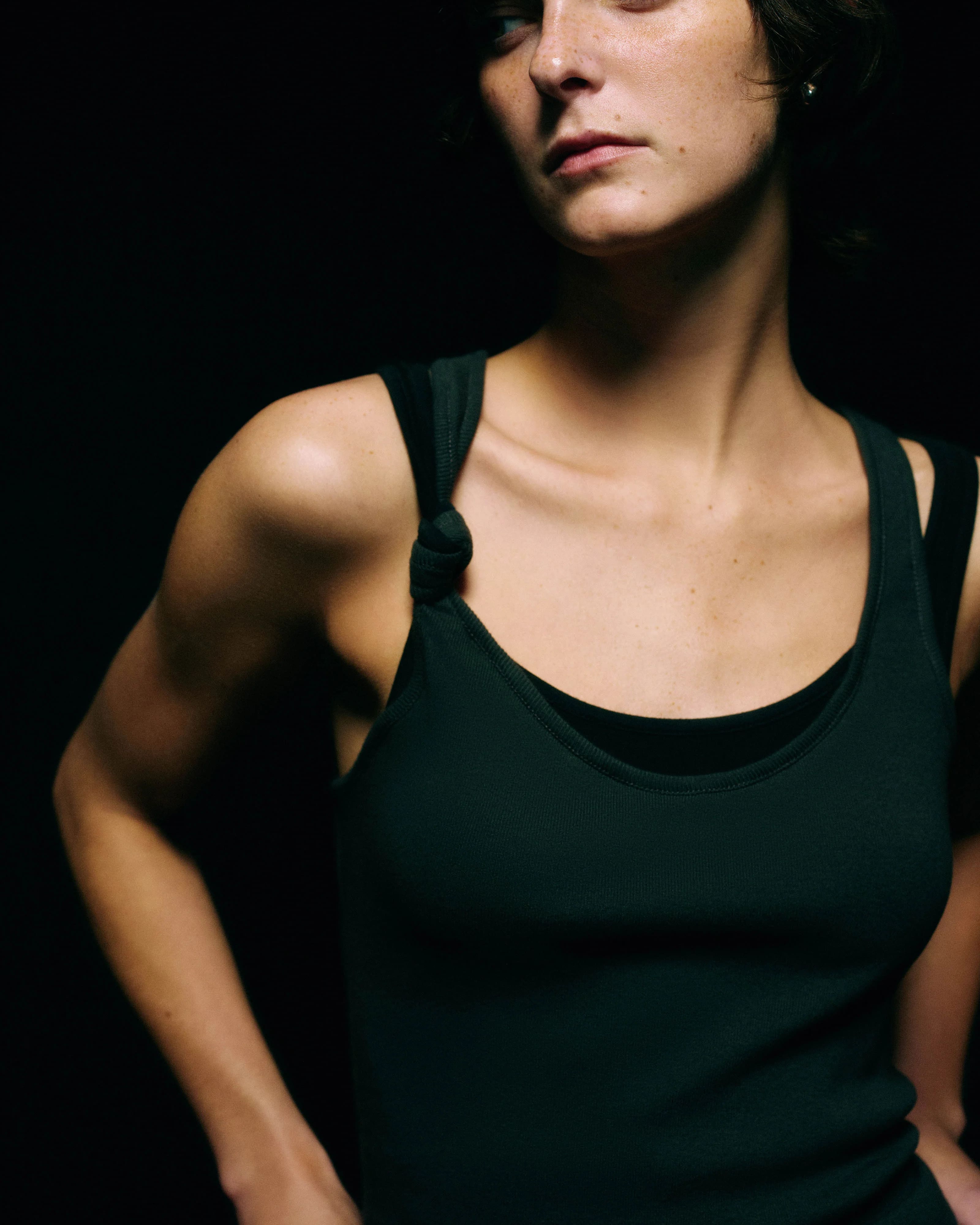 Close-up of the knotted double-strap shoulder detail on a black ribbed tank top against a black background