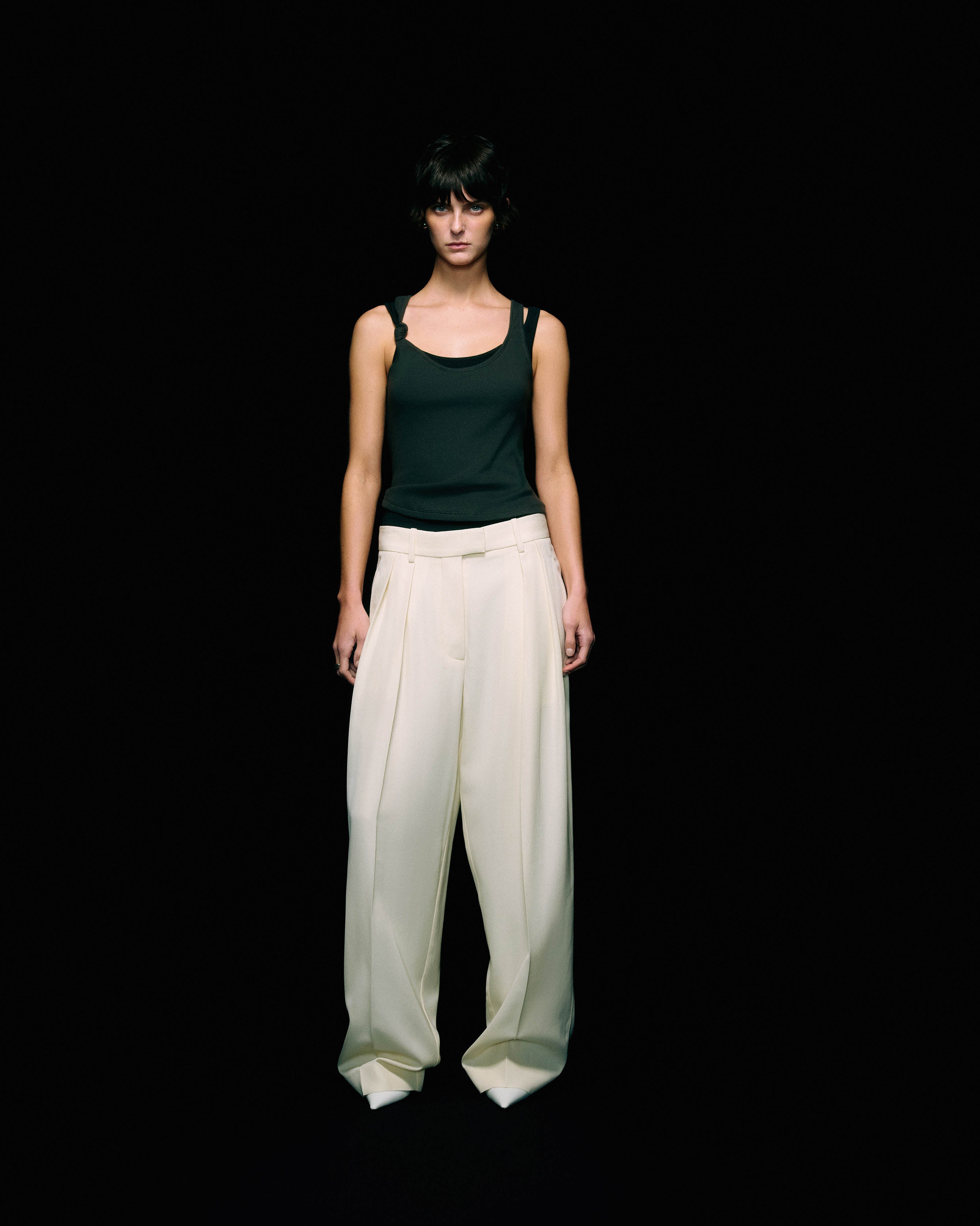 Model wearing a black ribbed double-strap tank top with wide-leg cream trousers, full-length front view against a black background