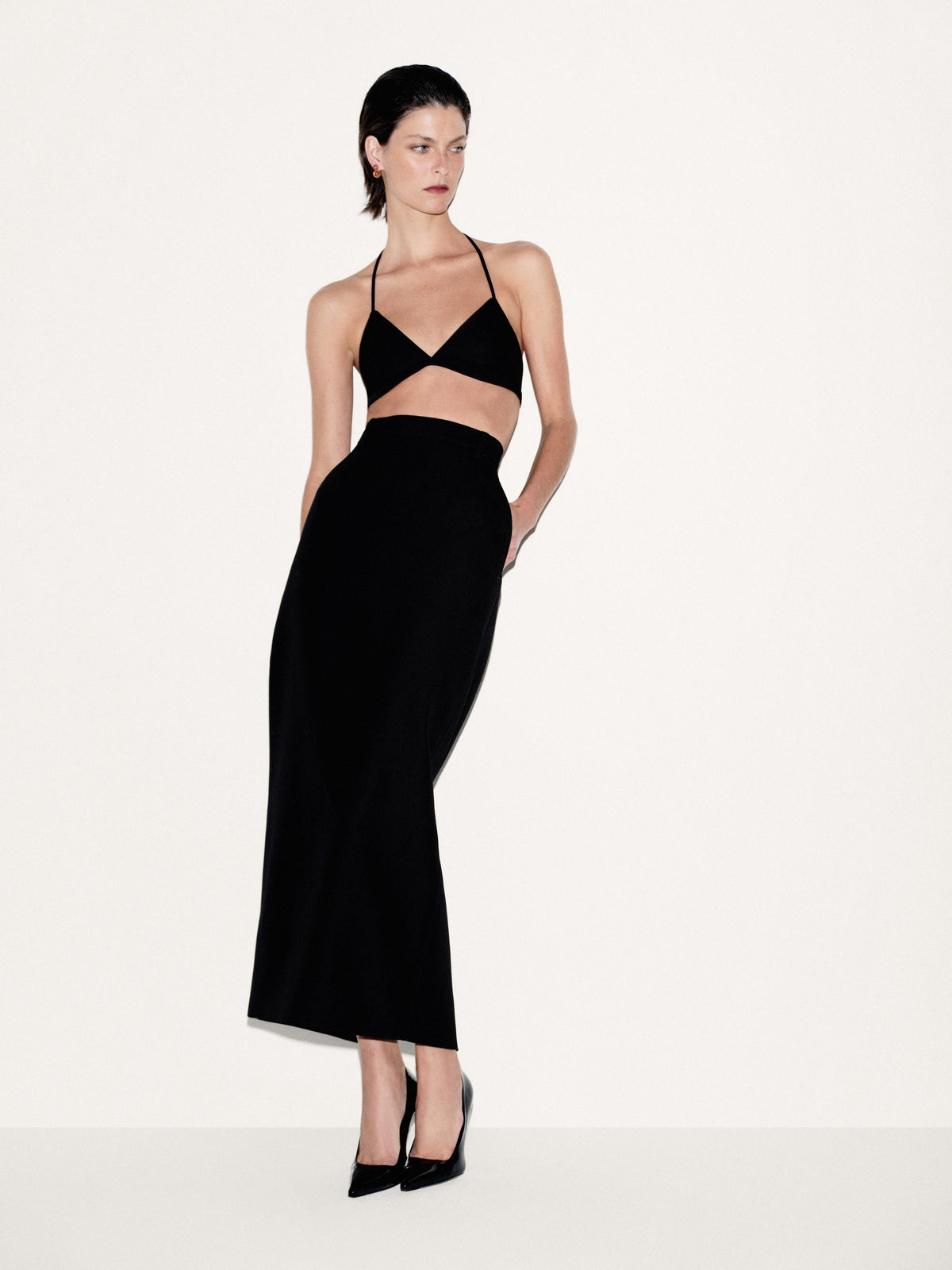 A model wearing a black two-piece outfit consisting of a halter-neck bra top with a matching high-waisted skirt, paired with black pointed-toe heels, against a plain off-white studio background.