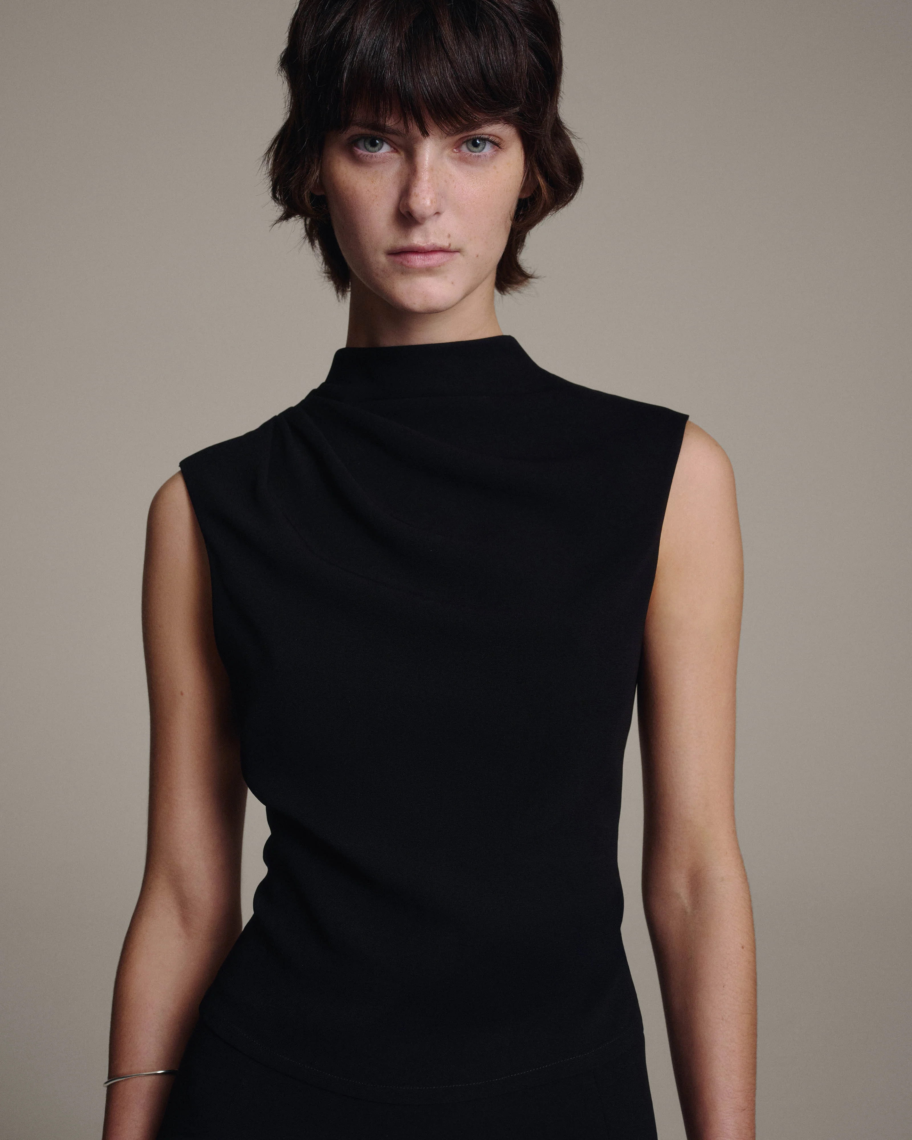 Model wearing a black sleeveless top with a softly draped high neckline, waist-up view against a taupe background