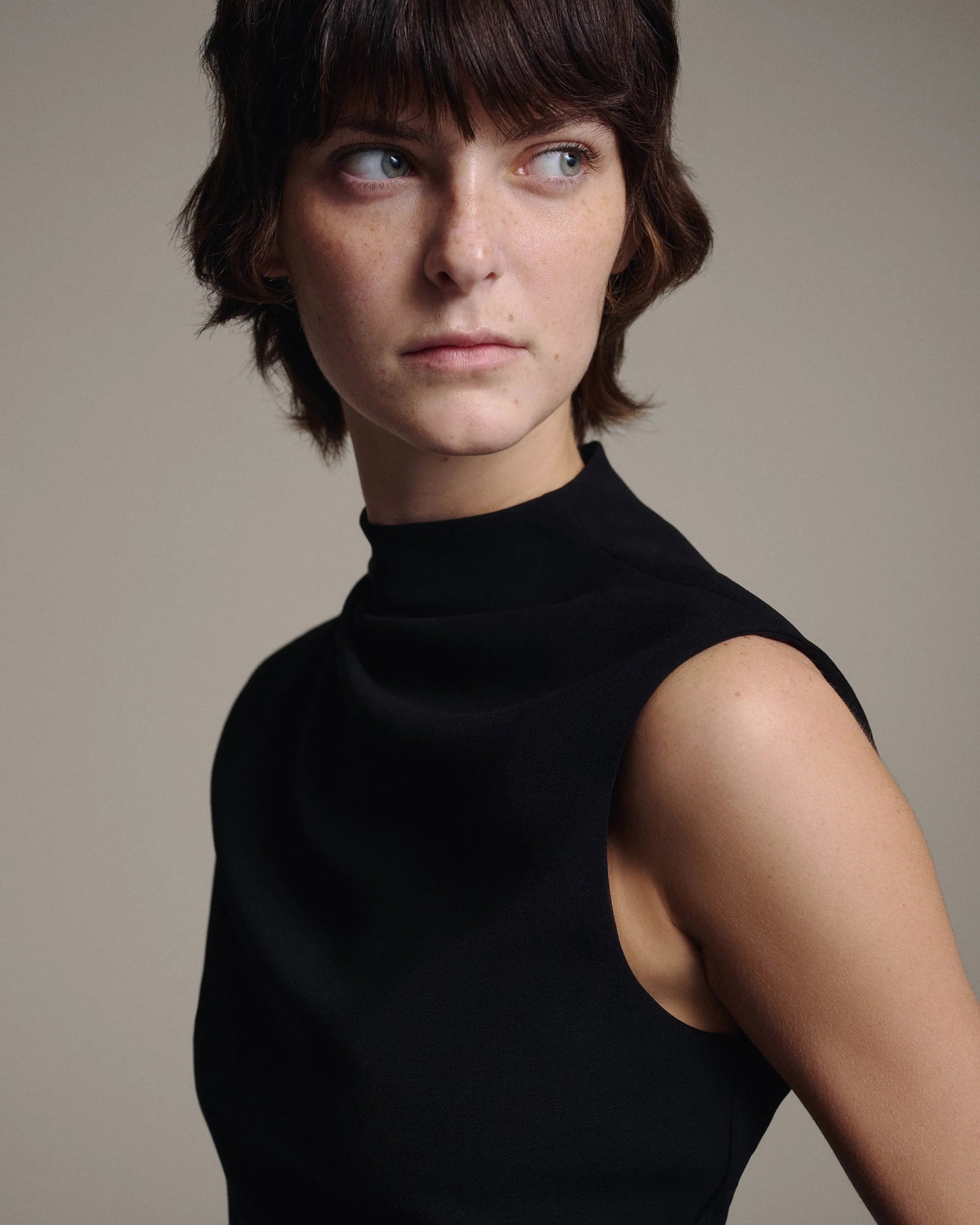 Model in a black sleeveless top with a softly draped high neckline, looking to the side, angled three-quarter portrait against a taupe background