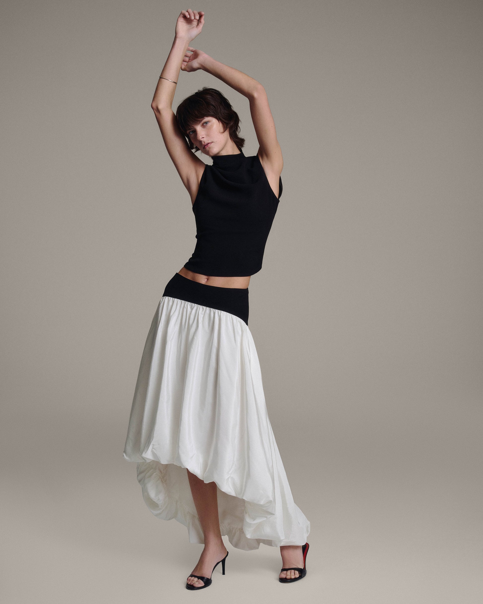 Model with arms raised overhead in a black sleeveless top and white bubble-hem skirt, exposing the midriff, dynamic full-length editorial pose against a taupe background