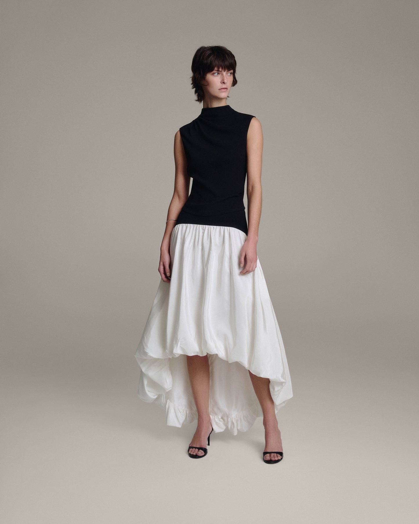 Model in a black sleeveless draped top and a white voluminous bubble skirt with a dramatic high-low hem, full-length view against a taupe background