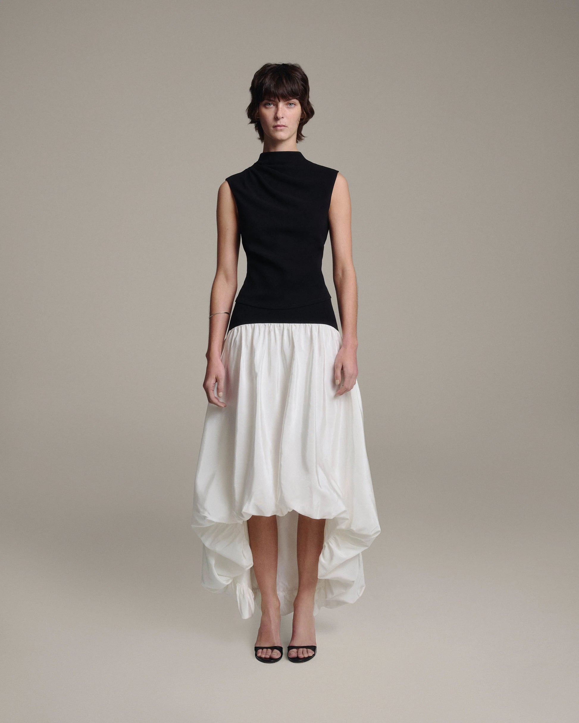 Model wearing a black sleeveless draped mock-neck top tucked into a white bubble-hem midi skirt with a high-low hem, full-length front view against a taupe background