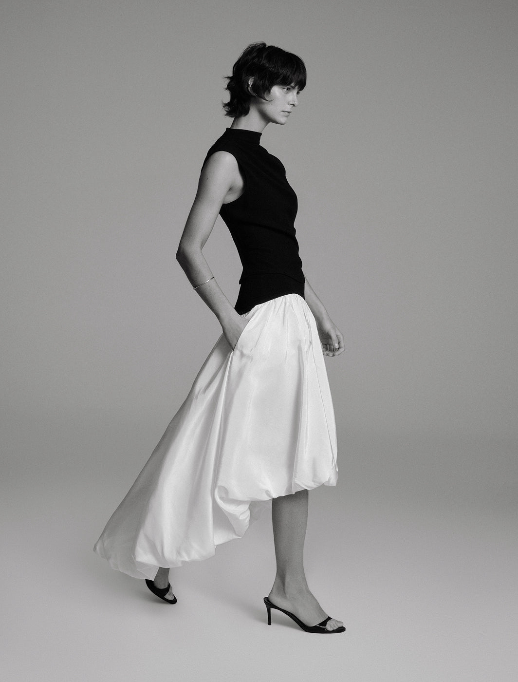 Black and white image of a side profile of a model walking in a black sleeveless draped top and white bubble-hem skirt with a trailing high-low hem against a taupe background
