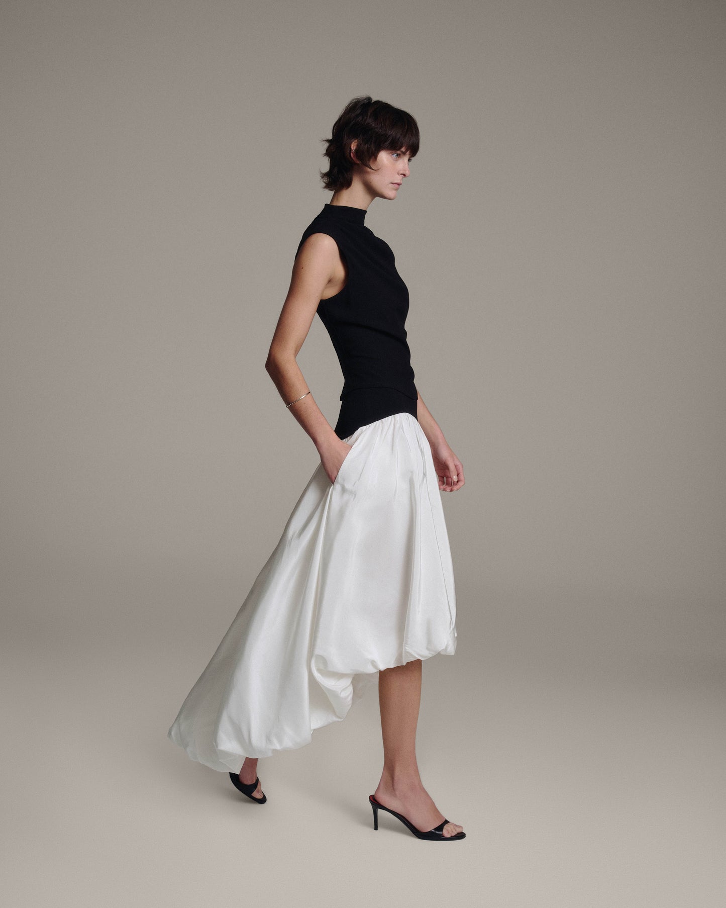 Side profile of a model walking in a black sleeveless draped top and white bubble-hem skirt with a trailing high-low hem against a taupe background