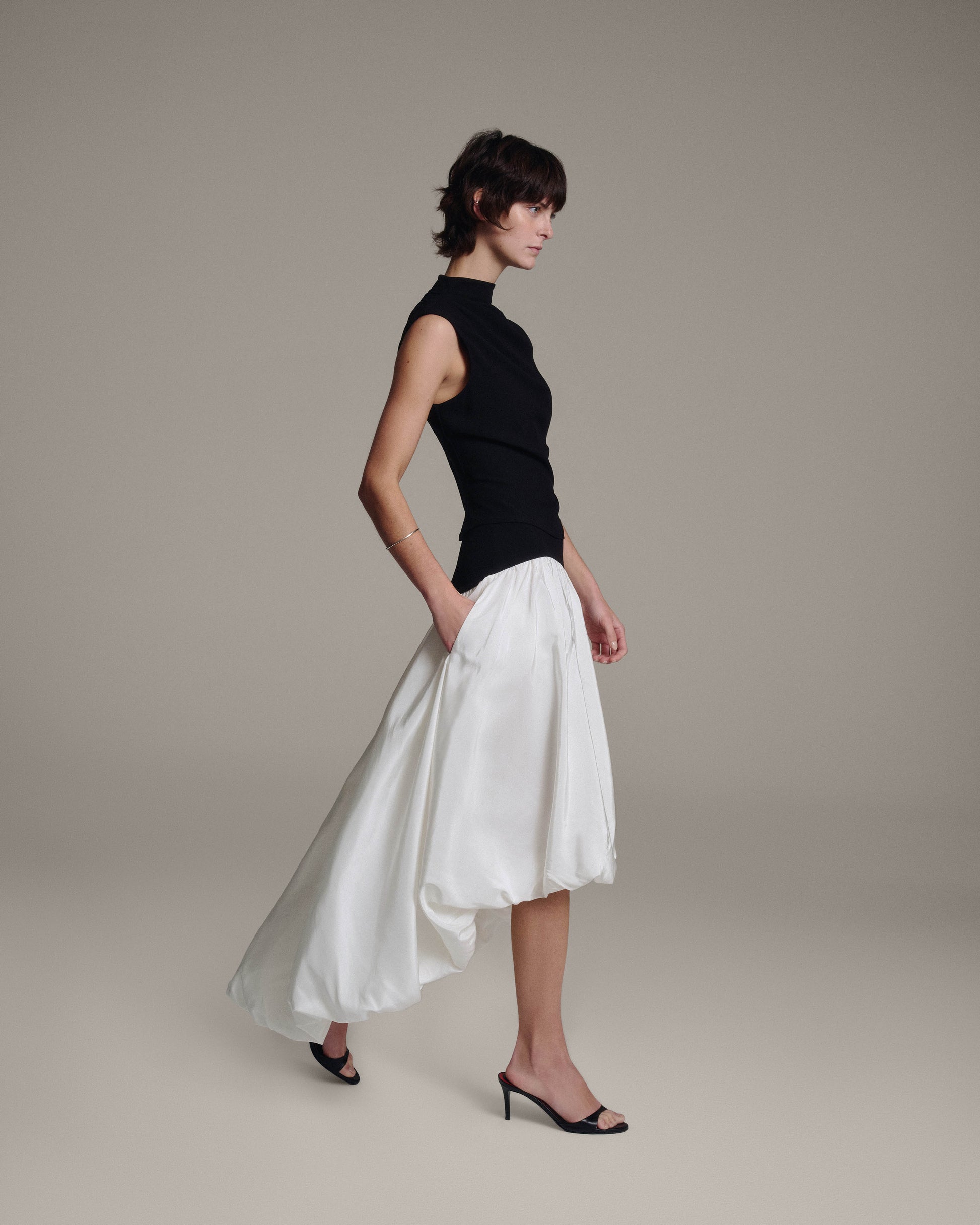 Side profile of a model walking in a black sleeveless draped top and white bubble-hem skirt with a trailing high-low hem against a taupe background
