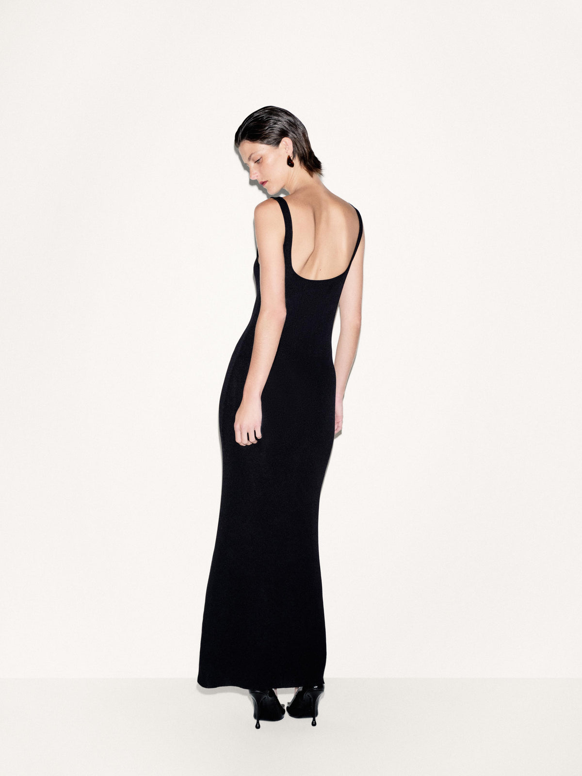 A woman modeling a sleeveless black maxi dress from the back, standing against a plain off-white studio background.