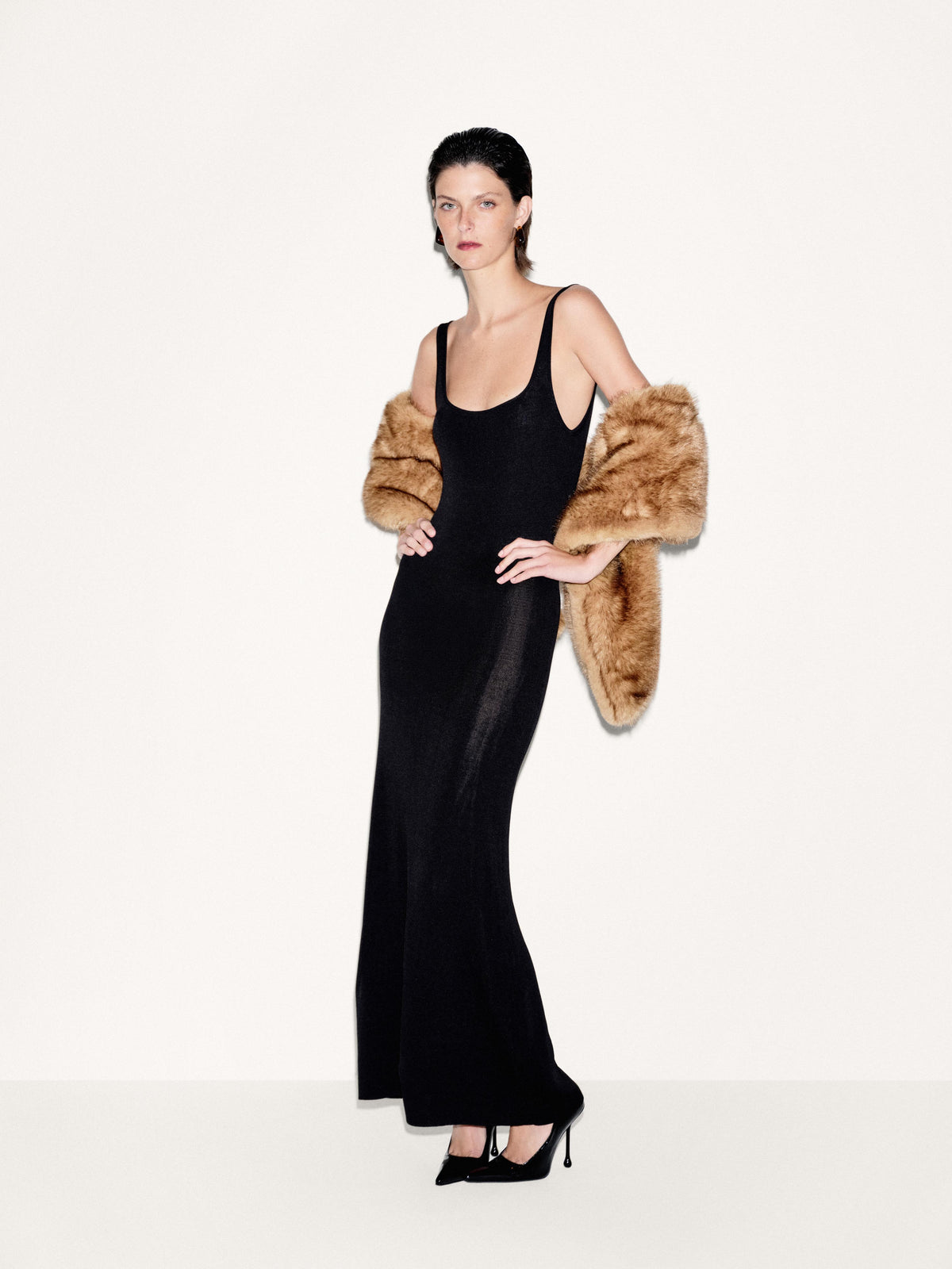 A model wearing a black sleeveless maxi dress with sheer side panels and a brown faux-fur cropped jacket, paired with black pointed-toe heels, against a plain off-white studio background.