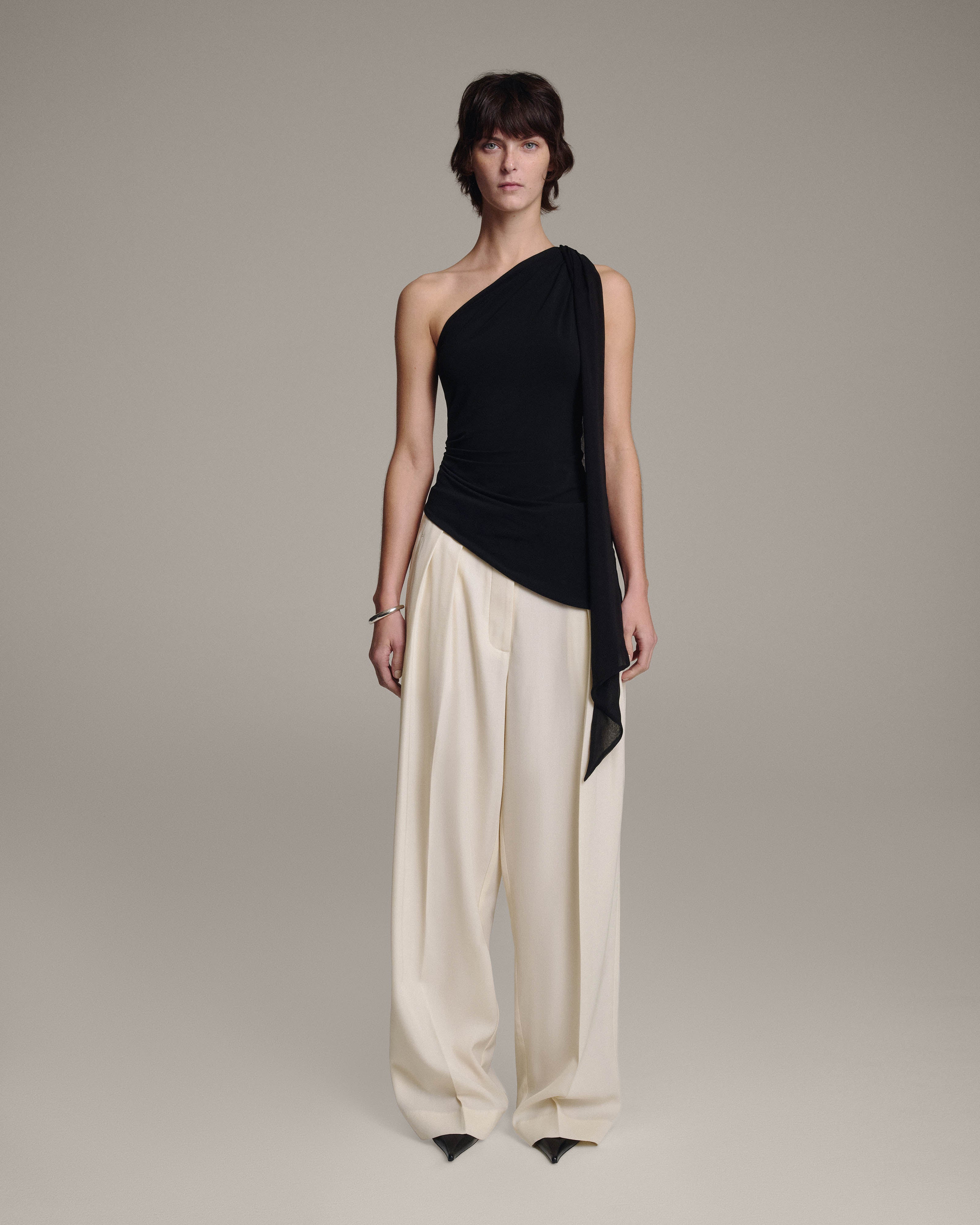 Model wearing a black one-shoulder top with a sheer chiffon shoulder tie draping down one side, paired with wide-leg cream trousers, full-length front view against a taupe background