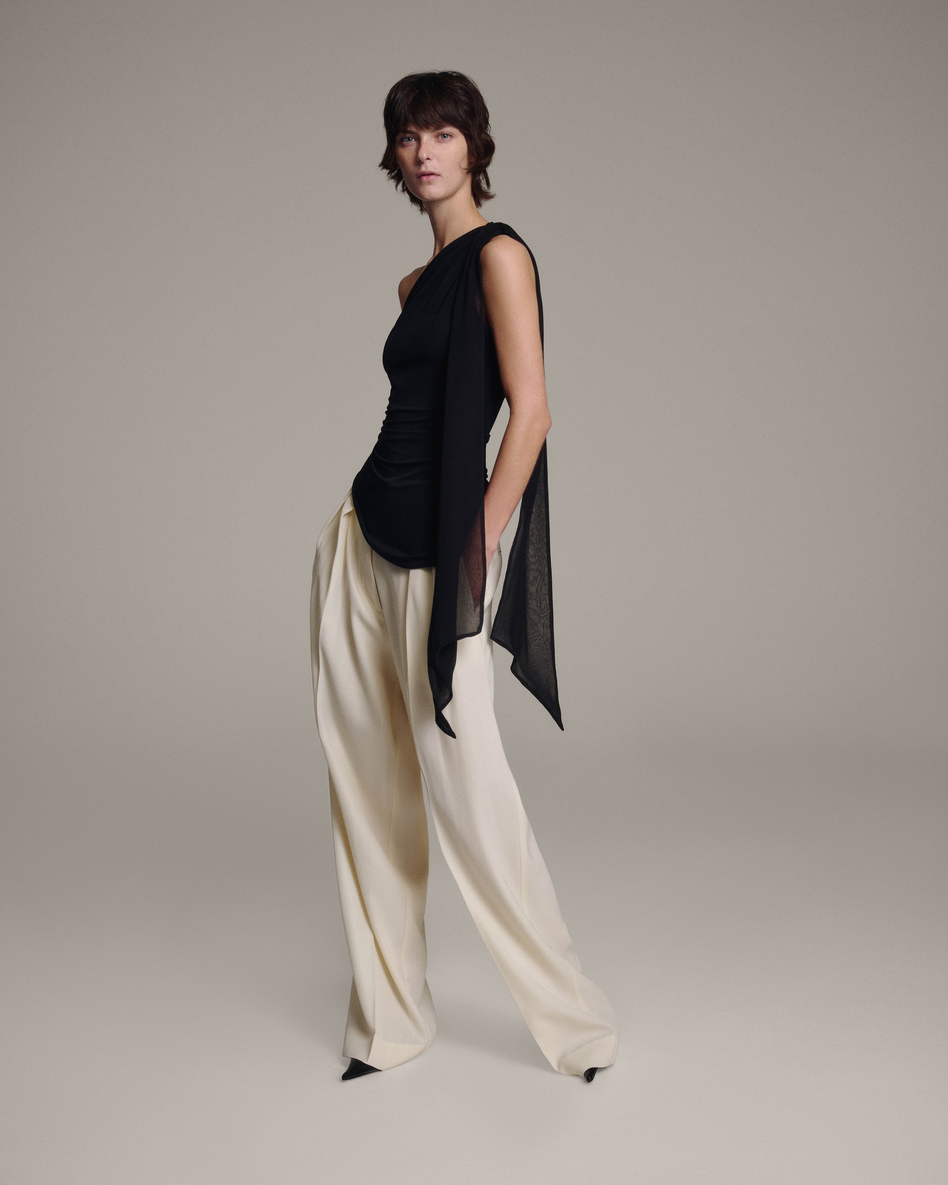 Model wearing a black one-shoulder top with a sheer chiffon shoulder tie cascading down one side, paired with high-waisted wide-leg cream trousers, full-length view against a taupe background
