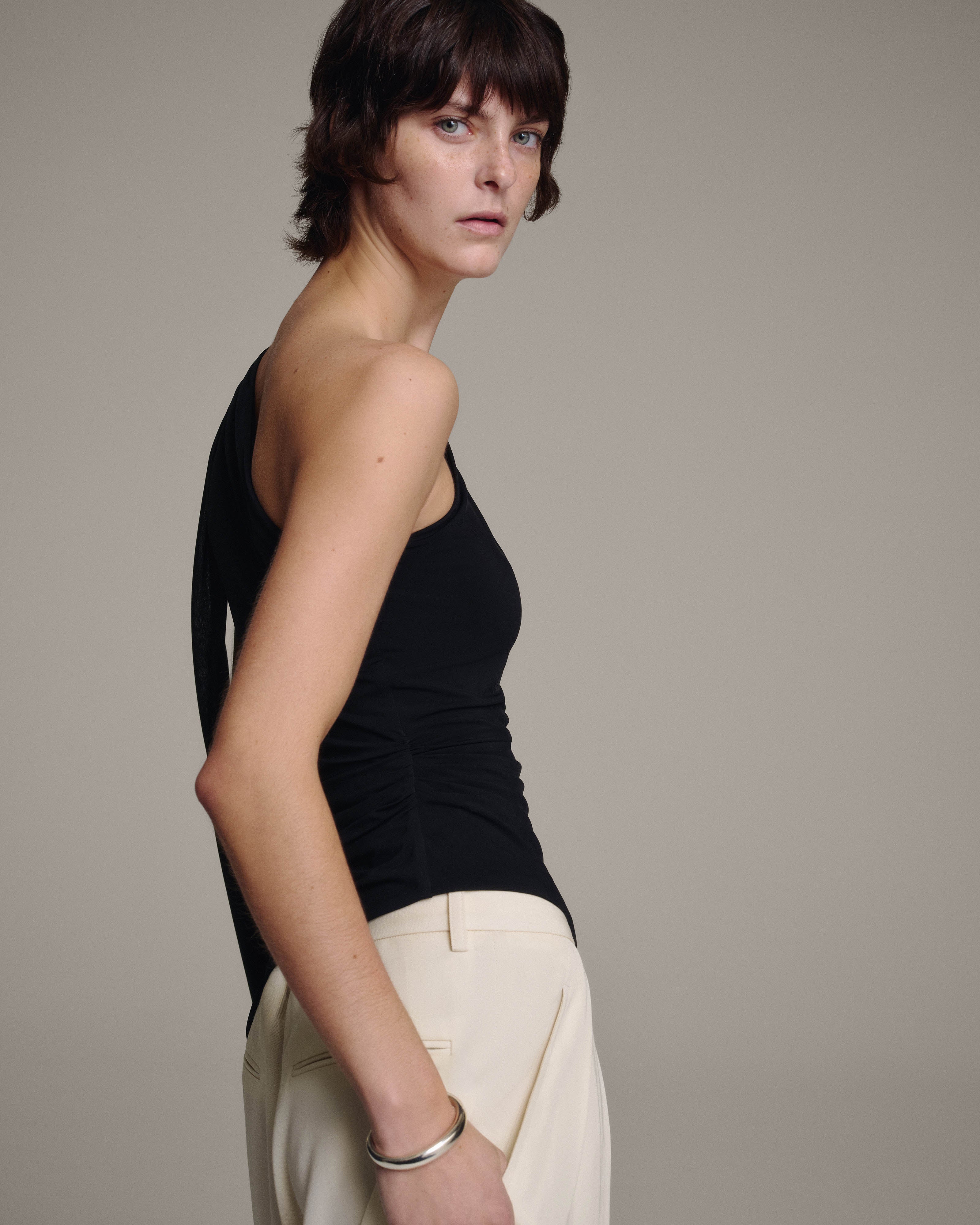 Side profile of a model in a black one-shoulder top with a sheer chiffon shoulder tie draping down the back, cream trousers at the waist, against a taupe background