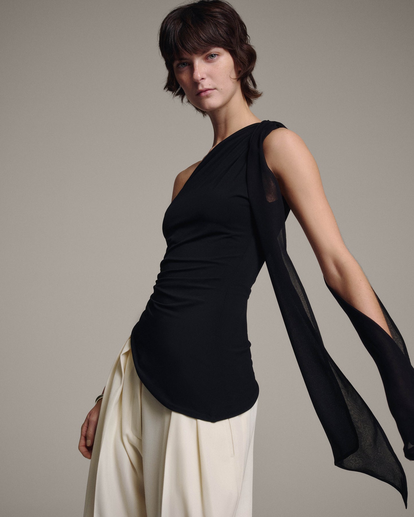 Model in a black one-shoulder top with a sheer chiffon tie trailing from the shoulder, angled three-quarter view against a taupe background