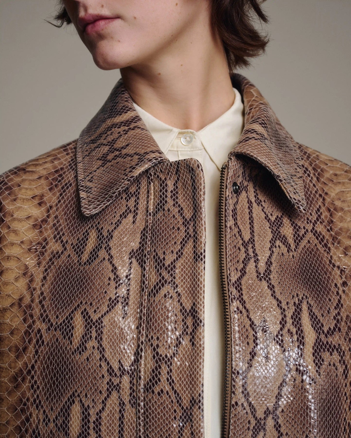 Close-up of a person wearing a brown and beige snakeskin-pattern leather jacket over a cream collared shirt, against a plain warm gray background.