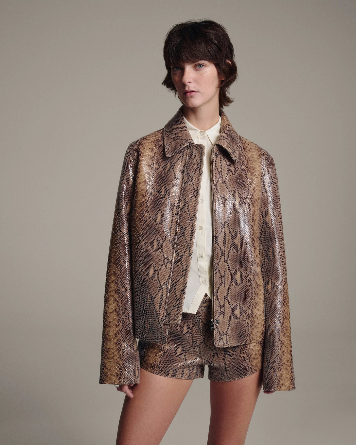 A model wearing a matching snakeskin-patterned leather jacket and shorts set in brown and beige tones, layered over a cream shirt, against a solid neutral studio background.