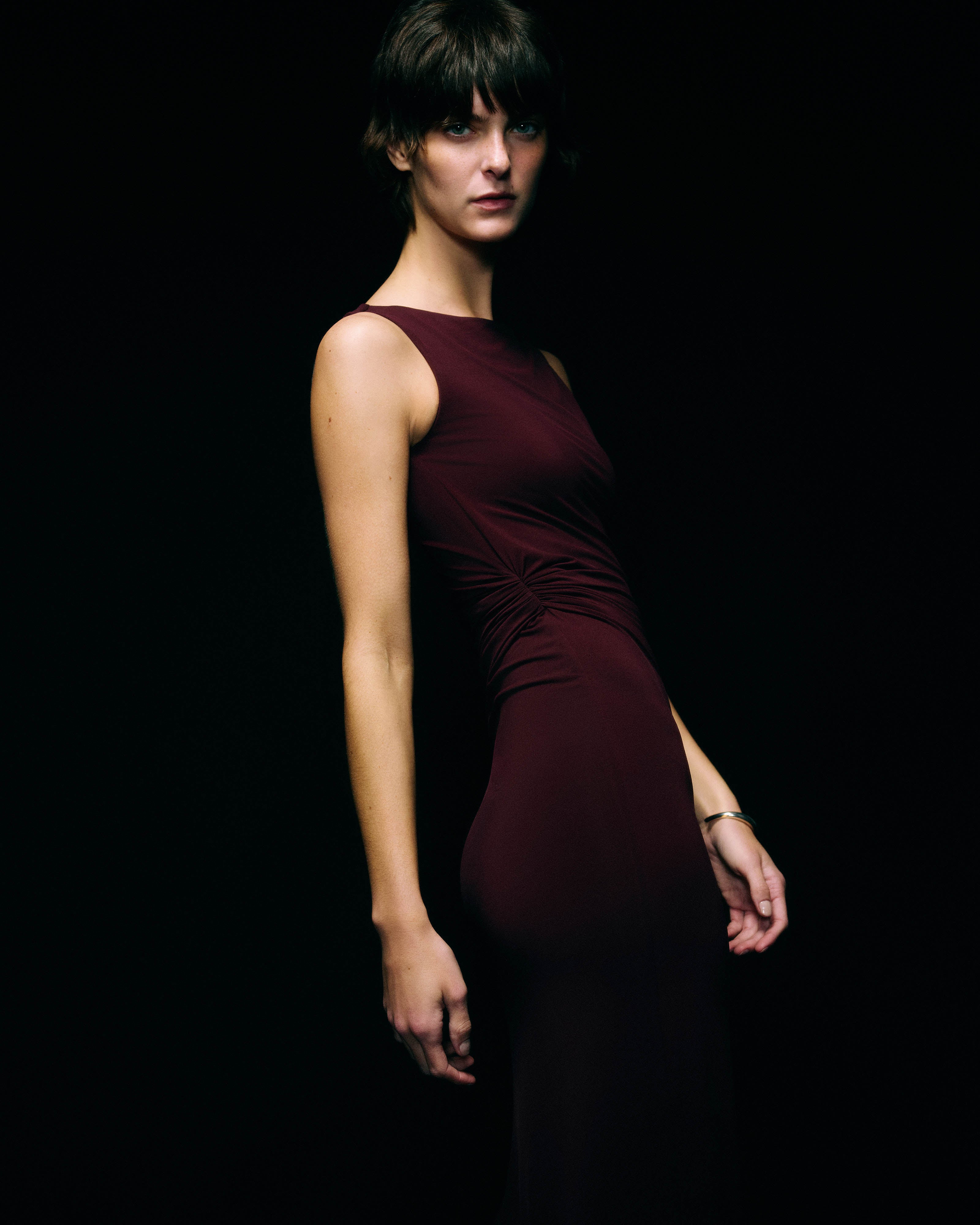 Side profile of a model in a fitted floor-length burgundy gown with a gathered knot at the waist and a high side slit against a black background
