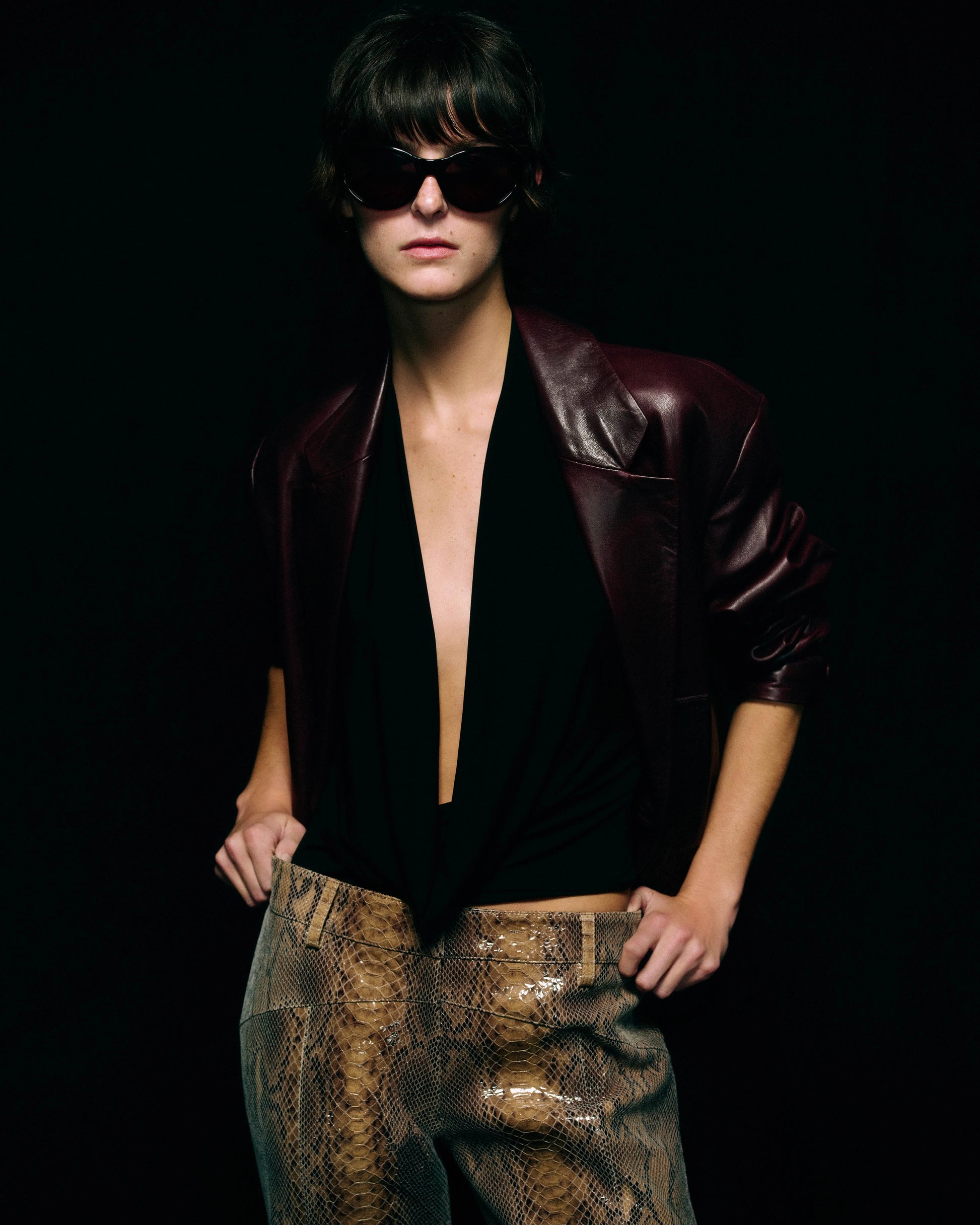 A model wearing a dark burgundy leather jacket, a black halter top, and snakeskin-patterned leather pants, set against a solid black background.