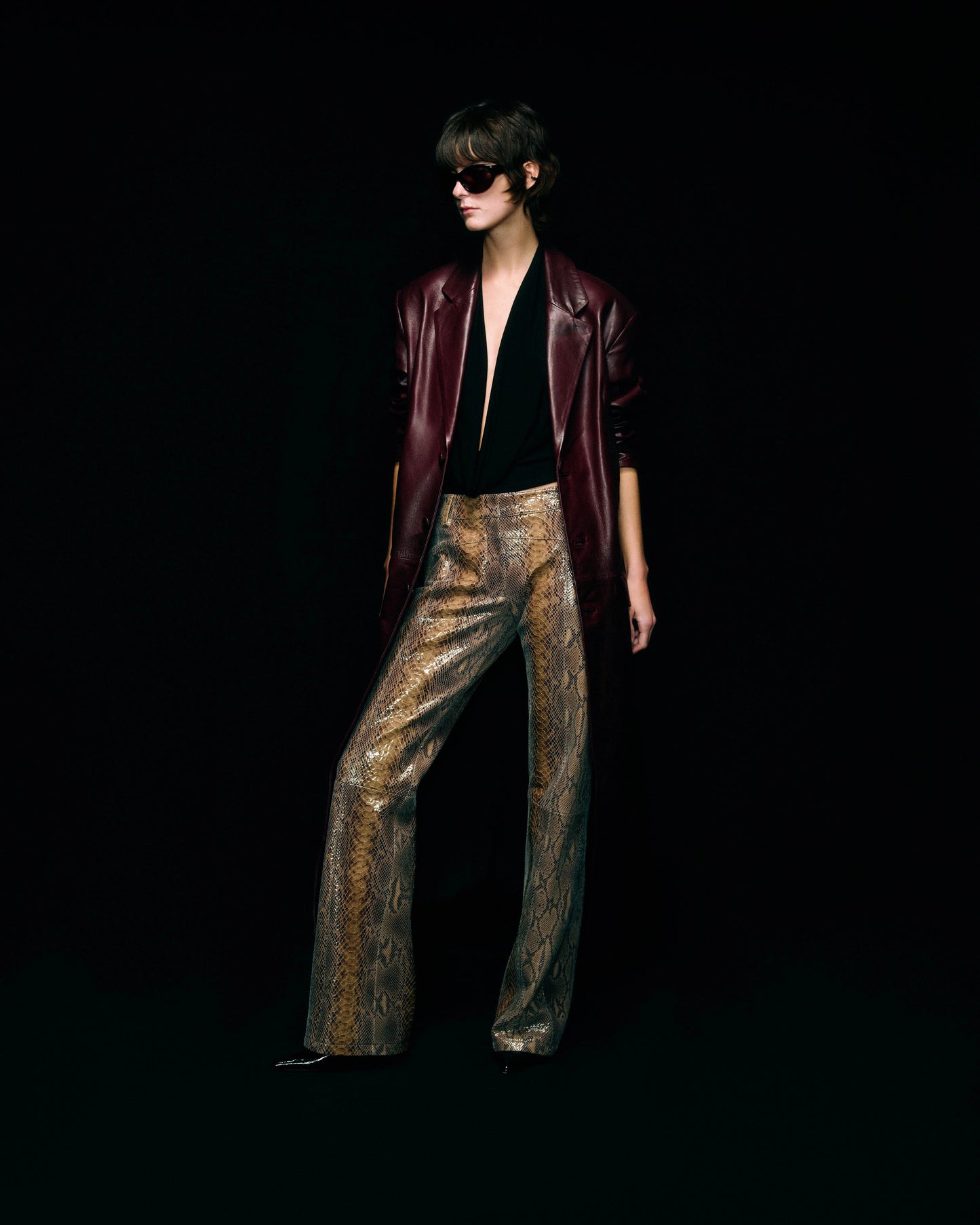 A model stands against a solid black background wearing a dark burgundy leather jacket, a black halter top, and snakeskin patterned leather trousers.