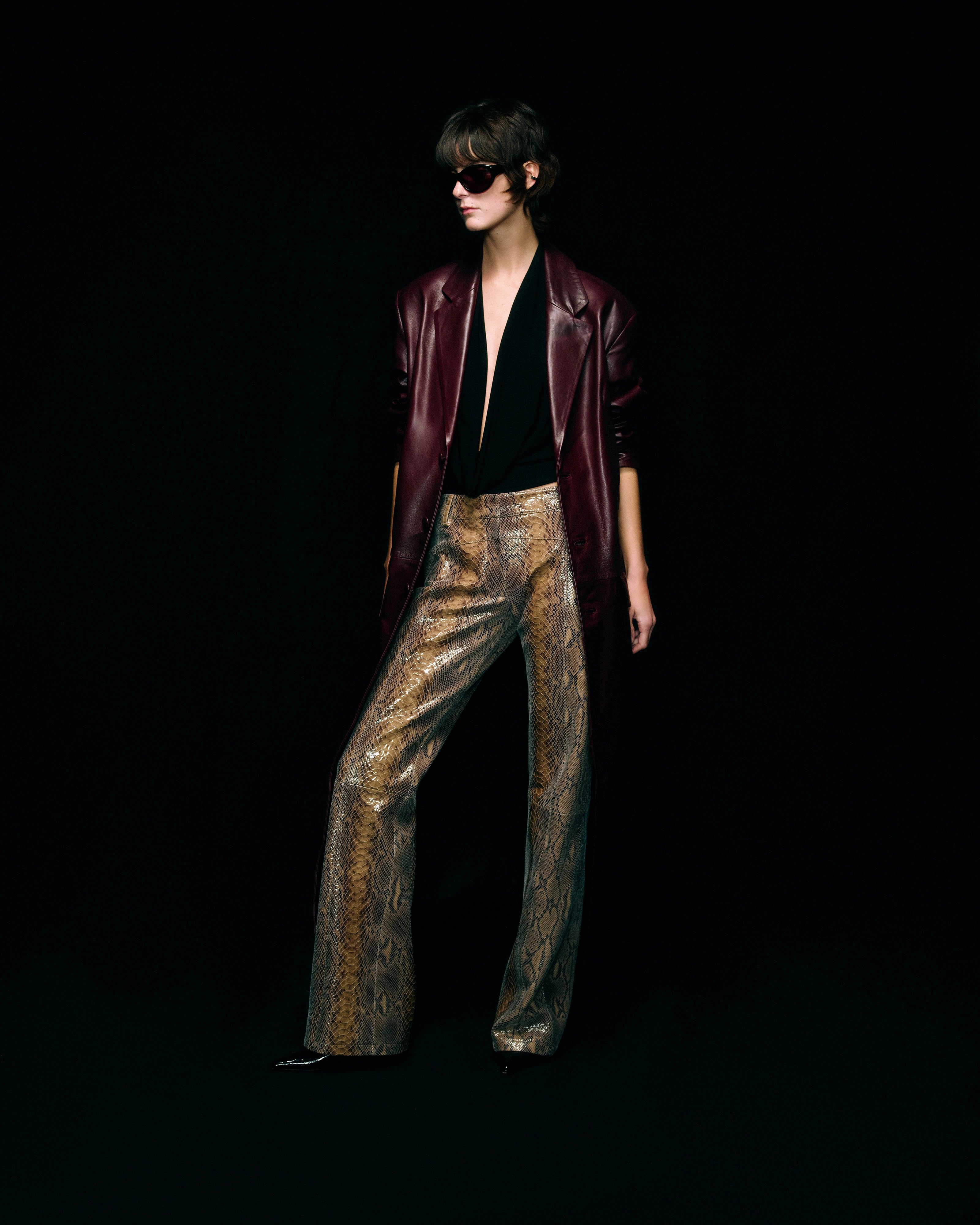 A model stands against a solid black background wearing a dark burgundy leather jacket, a black halter top, and snakeskin patterned leather trousers.