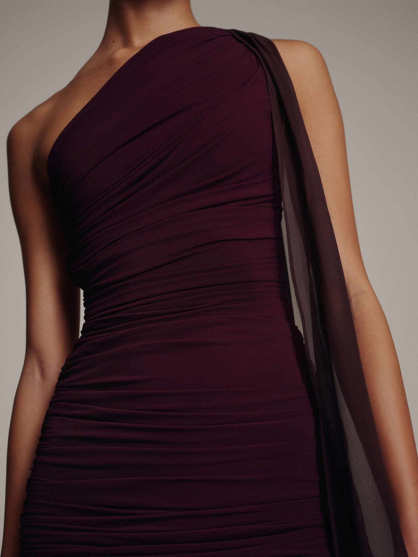 Close-up of the diagonal ruching across the bodice of a burgundy one-shoulder dress with a sheer chiffon shoulder tie against a taupe background
