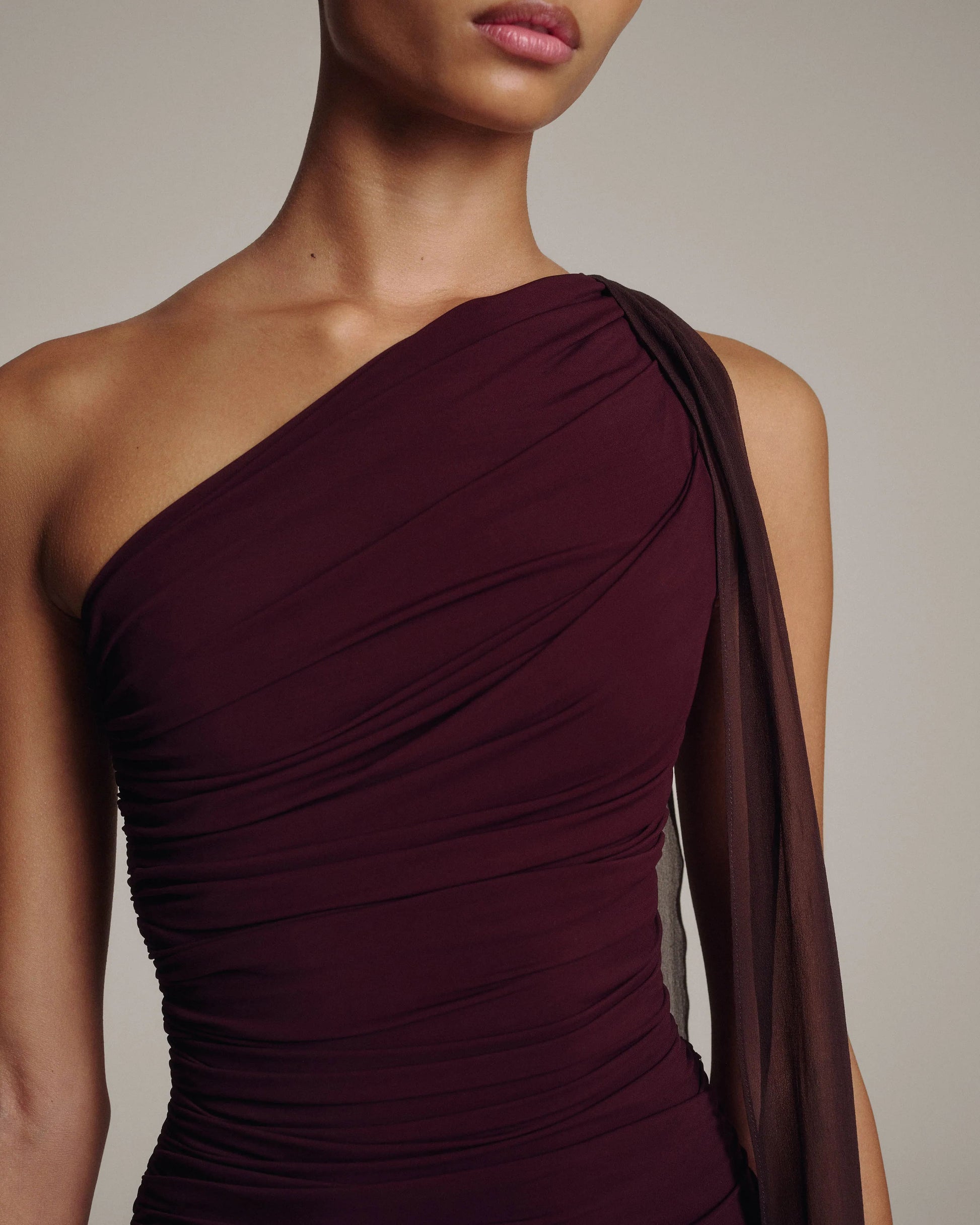 Model wearing a burgundy ruched one-shoulder dress with a draped neckline and sheer chiffon sash at the shoulder, waist-up view against a taupe background