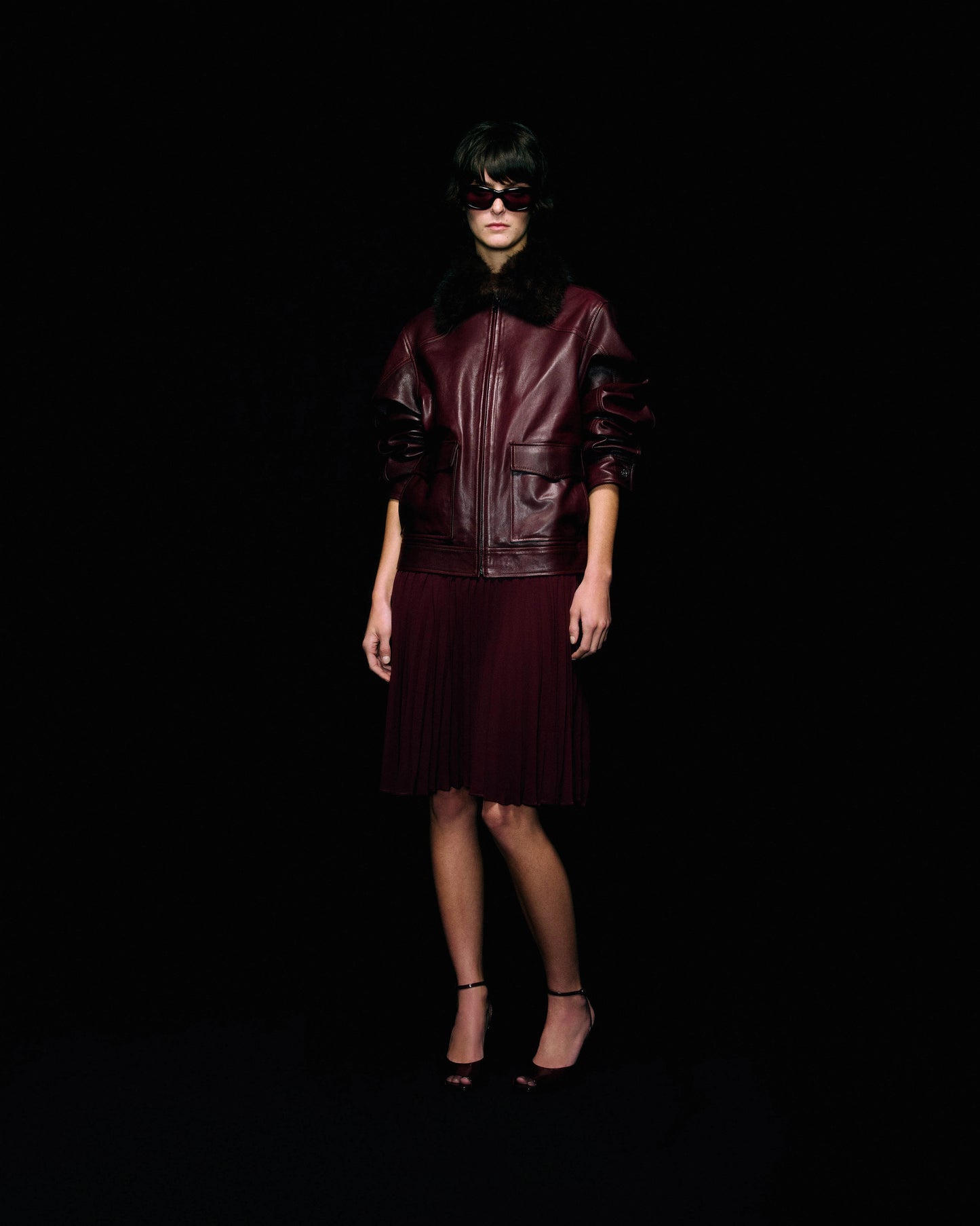 A model stands against a solid black background wearing a dark burgundy leather jacket with a black fur collar, a matching burgundy pleated skirt, and black high heels.