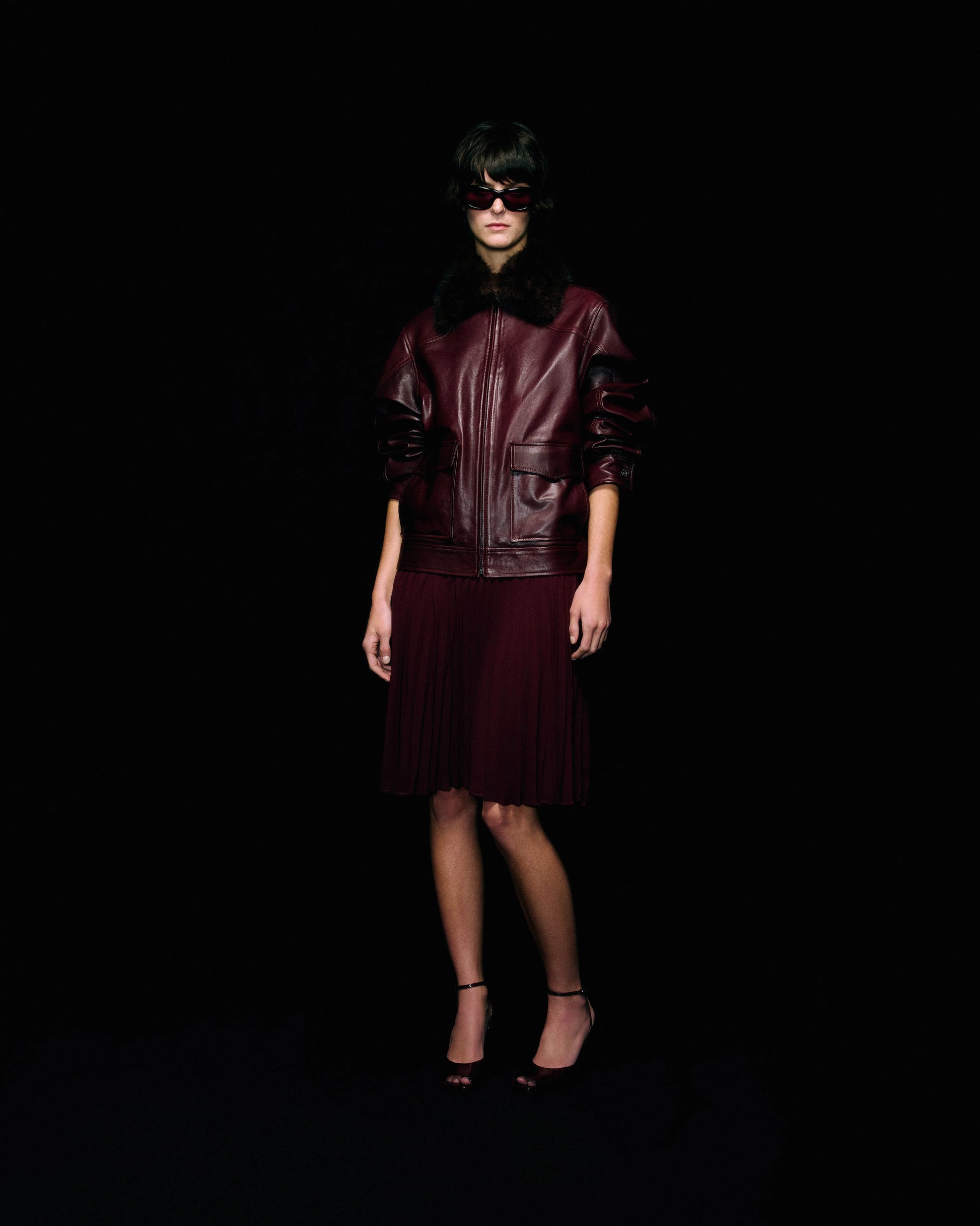 A model stands against a solid black background wearing a dark burgundy leather jacket with a black fur collar, a matching burgundy pleated skirt, and black high heels.
