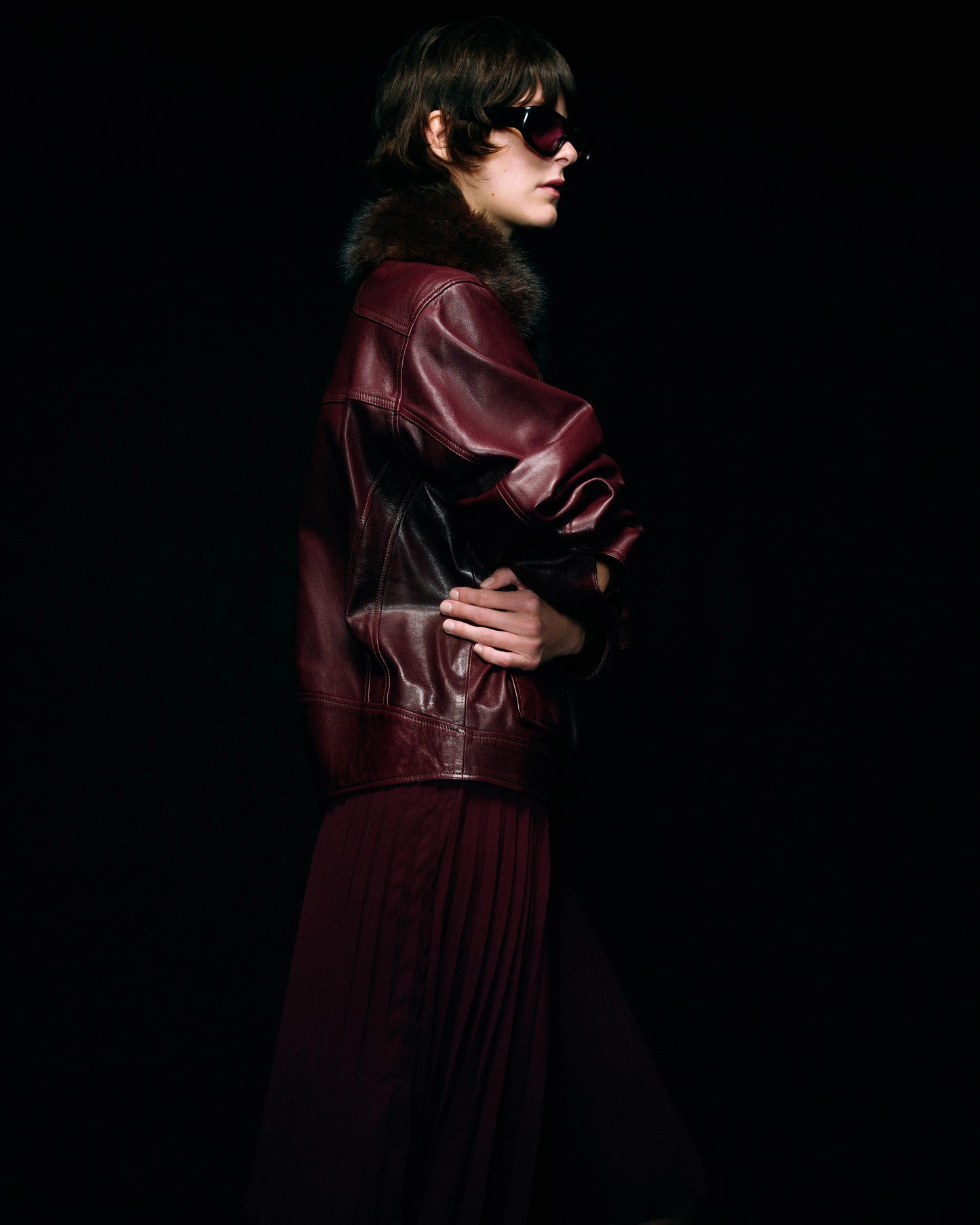 A model in profile wearing a burgundy leather jacket with a dark fur collar, paired with a matching pleated skirt, set against a solid black background.