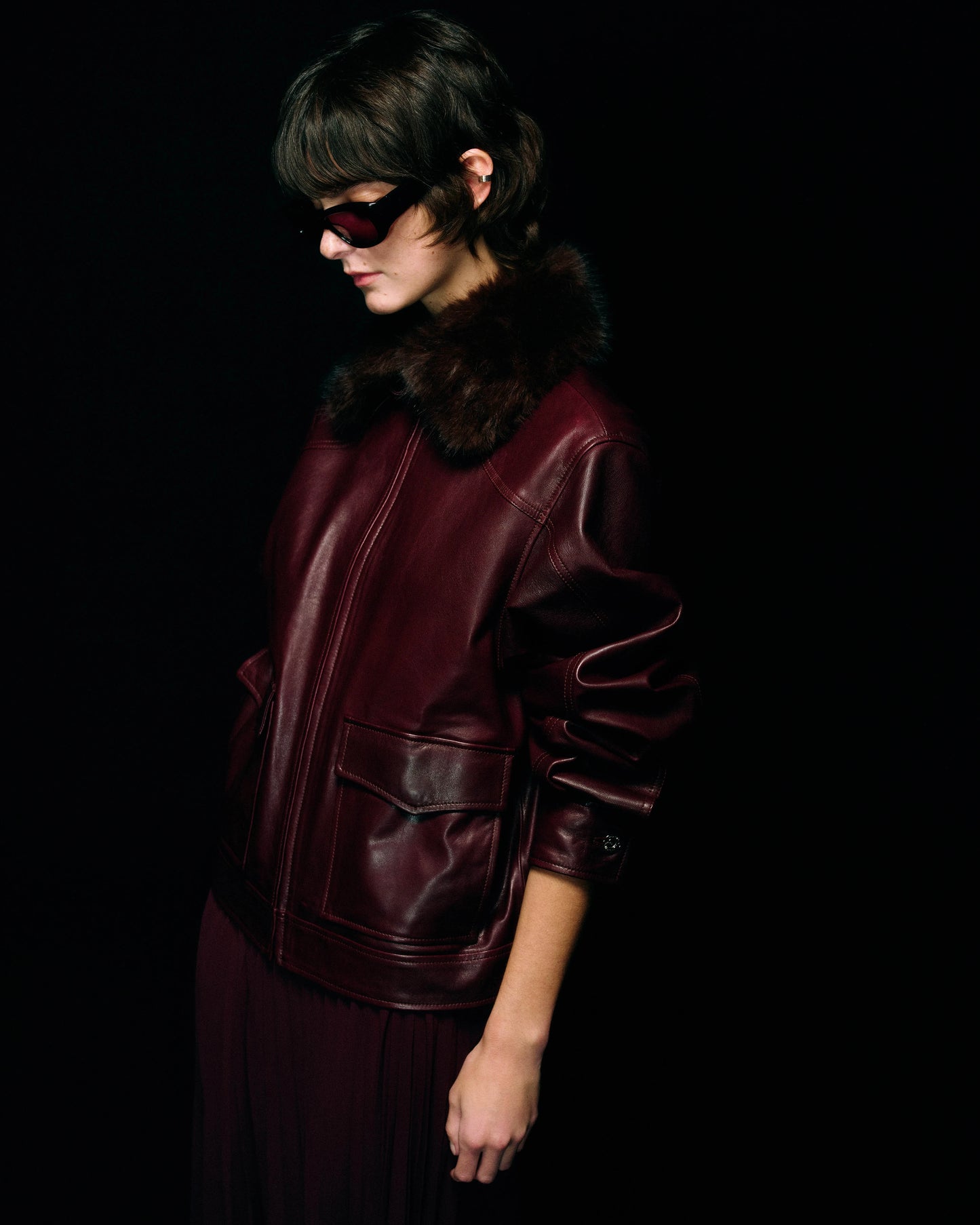 A model in profile wearing a dark burgundy leather jacket with a dark brown fur collar, black sunglasses, and a matching burgundy pleated skirt against a solid black background.
