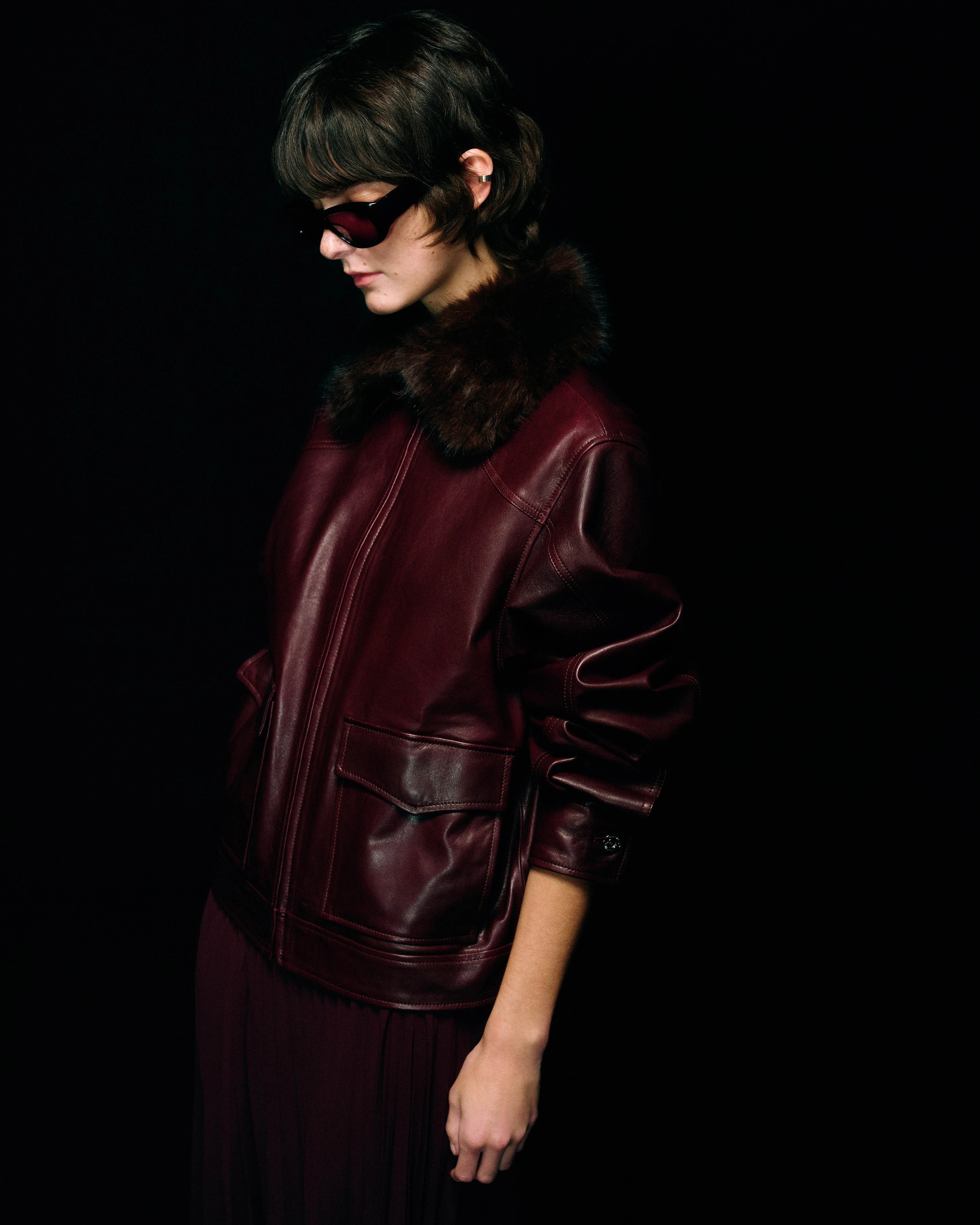 A model in profile wearing a dark burgundy leather jacket with a dark brown fur collar, black sunglasses, and a matching burgundy pleated skirt against a solid black background.