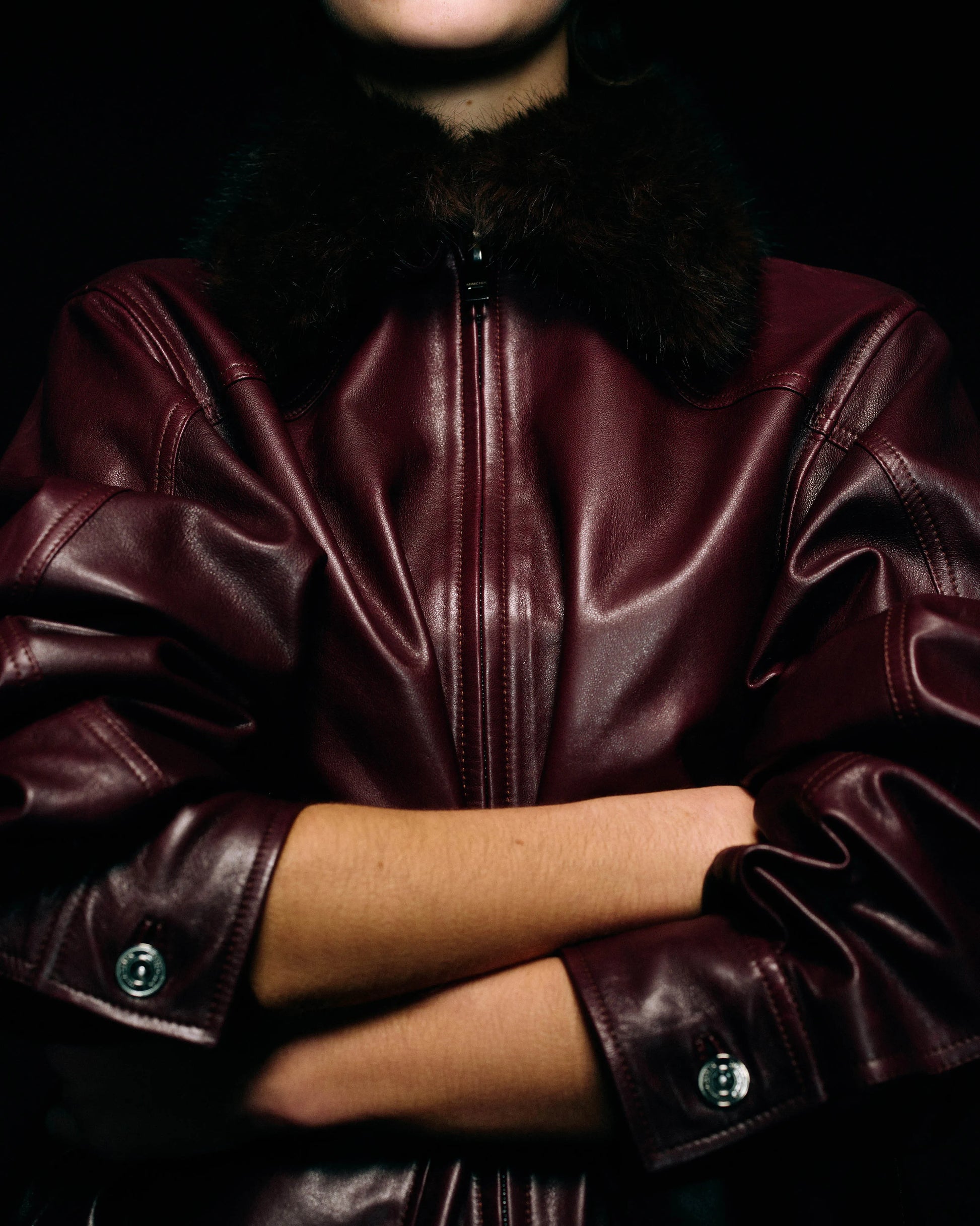 A close-up of a person wearing a dark burgundy leather jacket with a dark brown fur collar, with arms crossed against a black background.