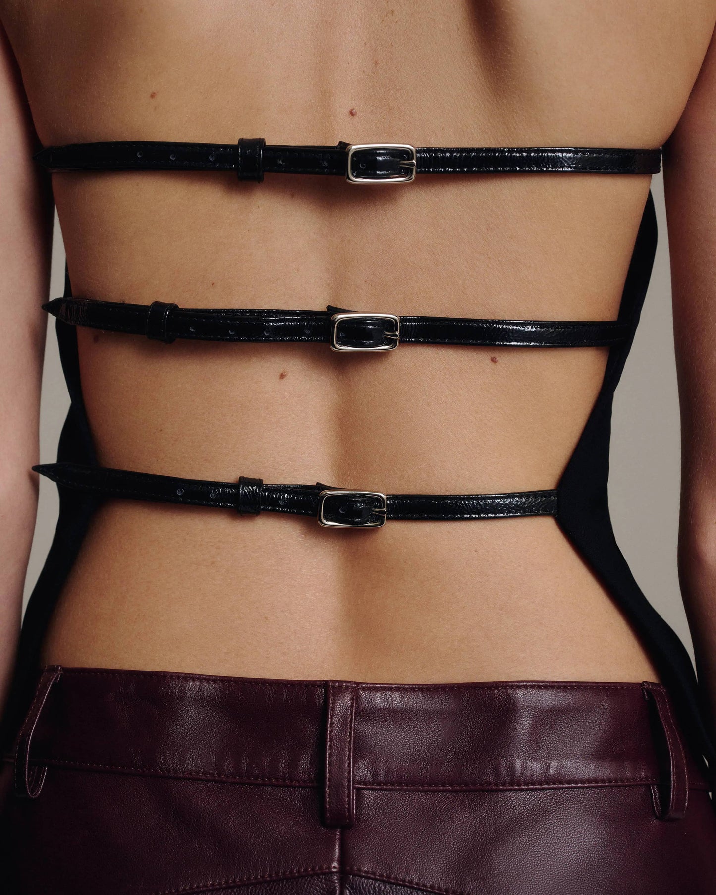 Close-up of the open back of a black halter top with three horizontal buckled leather straps, above the waistband of burgundy leather trousers, against a taupe background